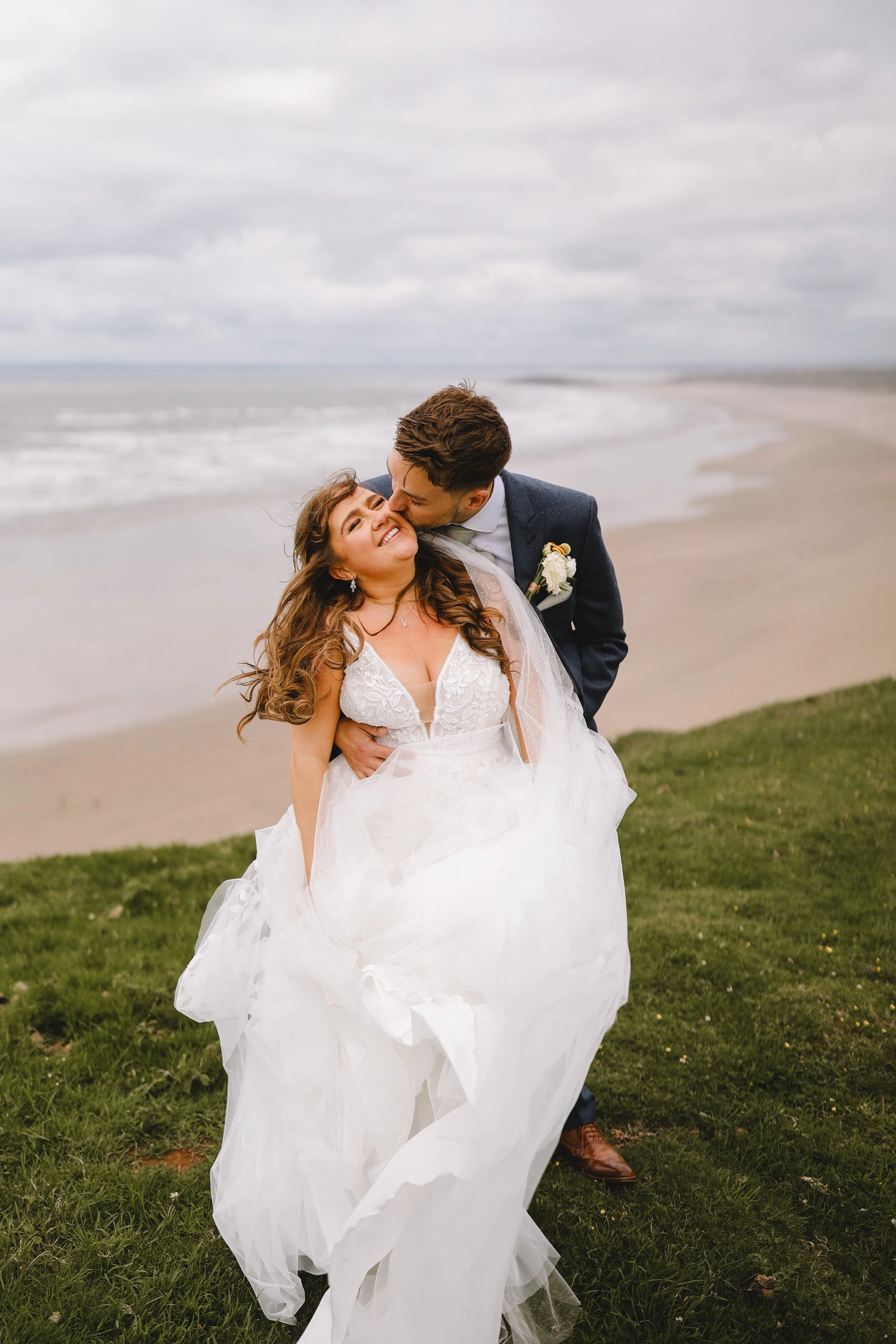A newlywed couple embracing on a seaside cliff, with the groom in a navy suit and the bride in a white wedding dress smiling joyfully. The ocean and beach are visible in the background under a cloudy sky.