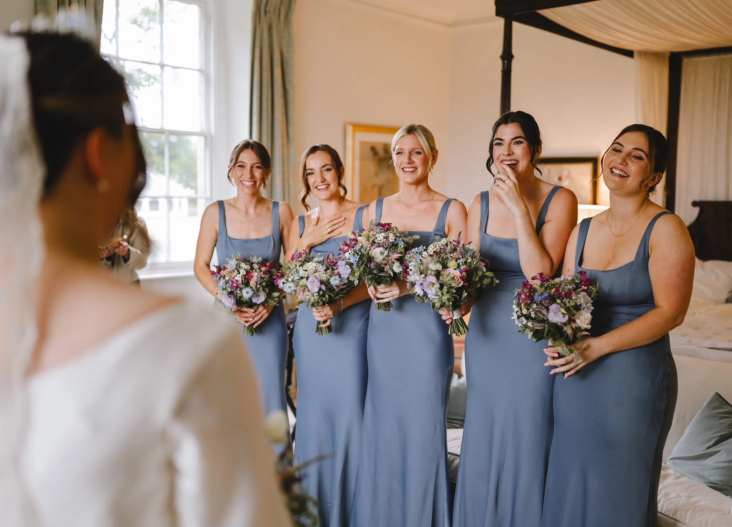 Bride facing bridesmaids wearing blue dresses holding bouquets, smiling indoors.