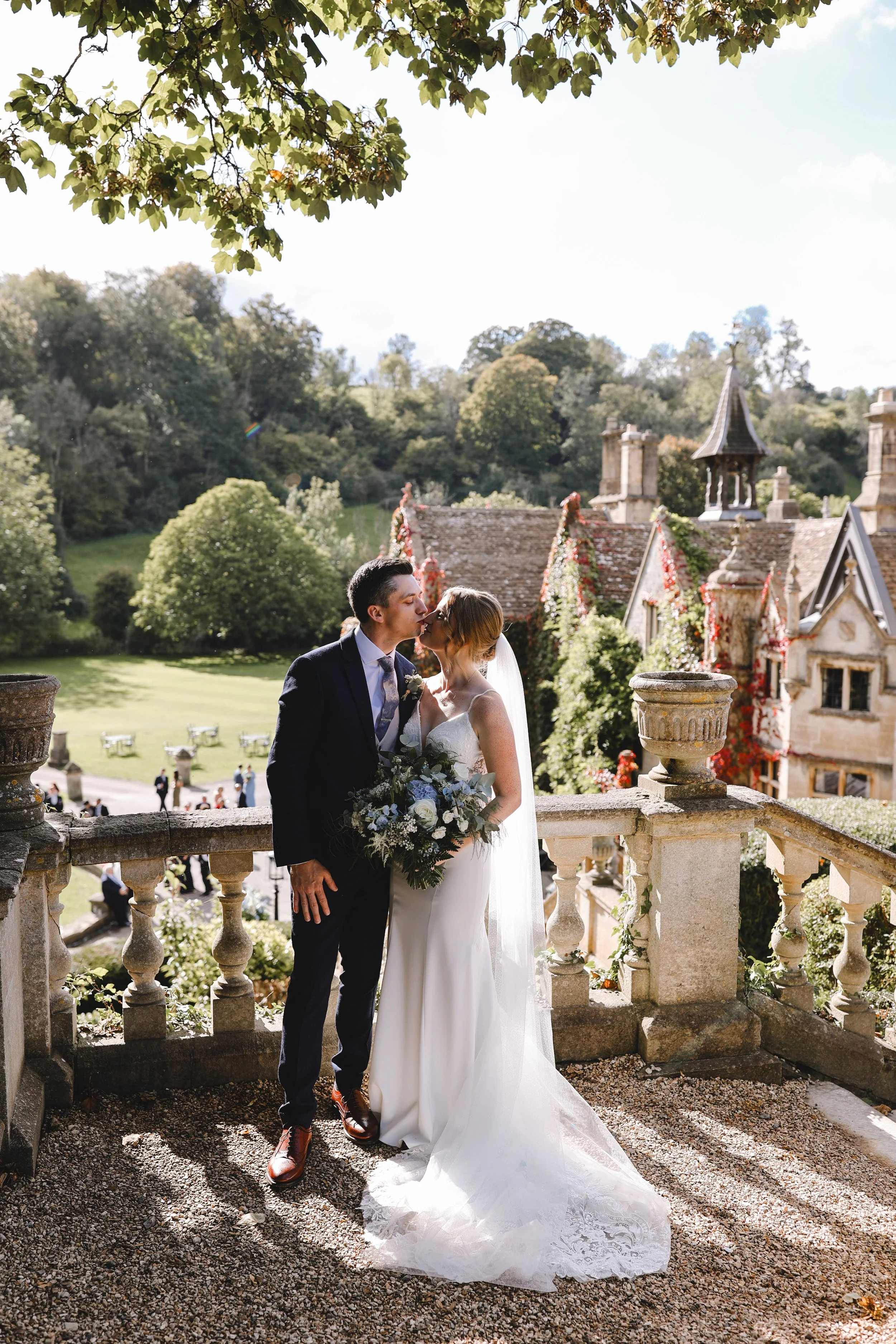 Bride and groom kissing on a sunny day, standing on a stone terrace with a scenic view of greenery and an old, vine-covered building in the background.