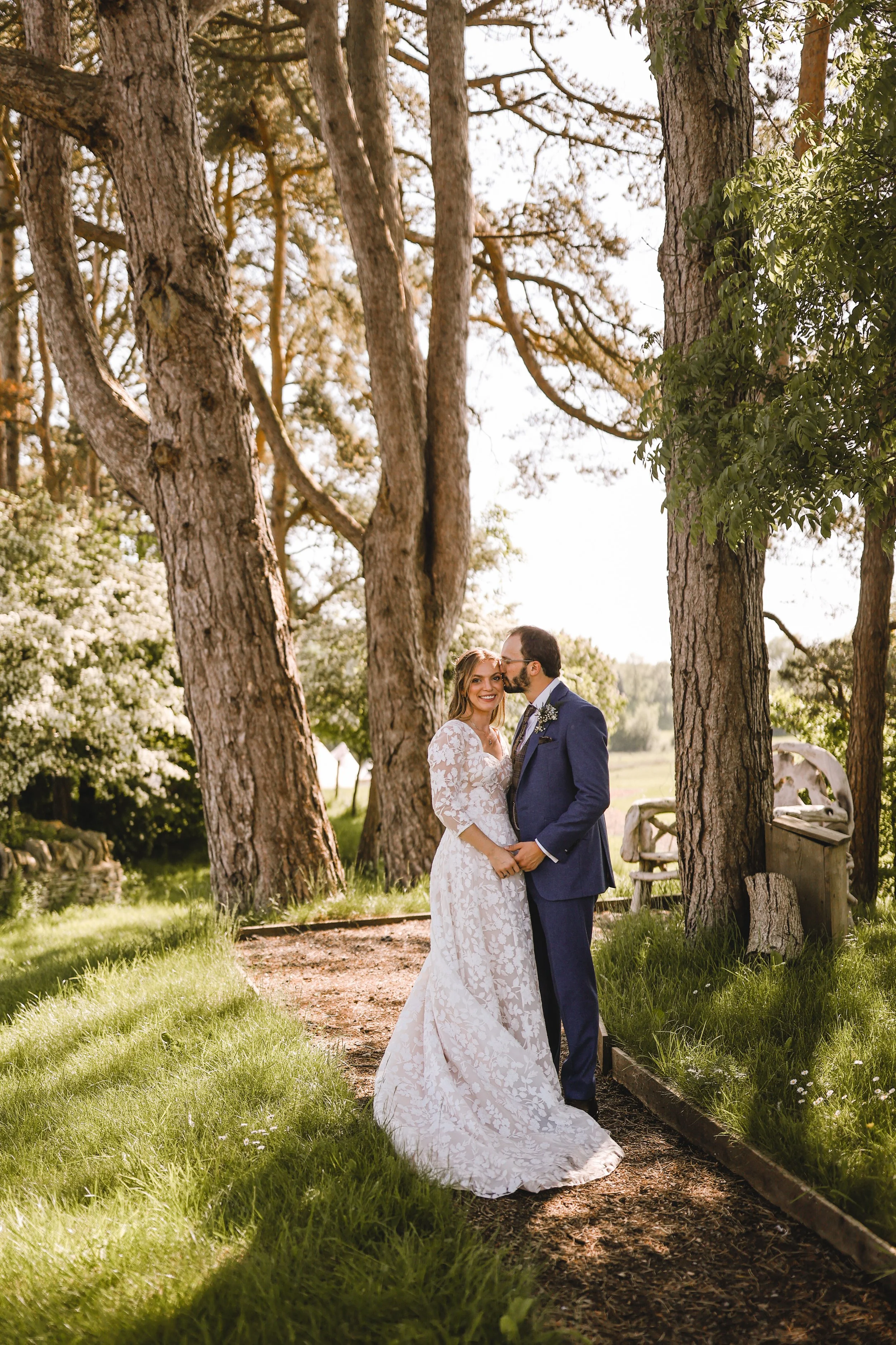 A couple in a wedding dress and suit stand outdoors surrounded by tall trees.