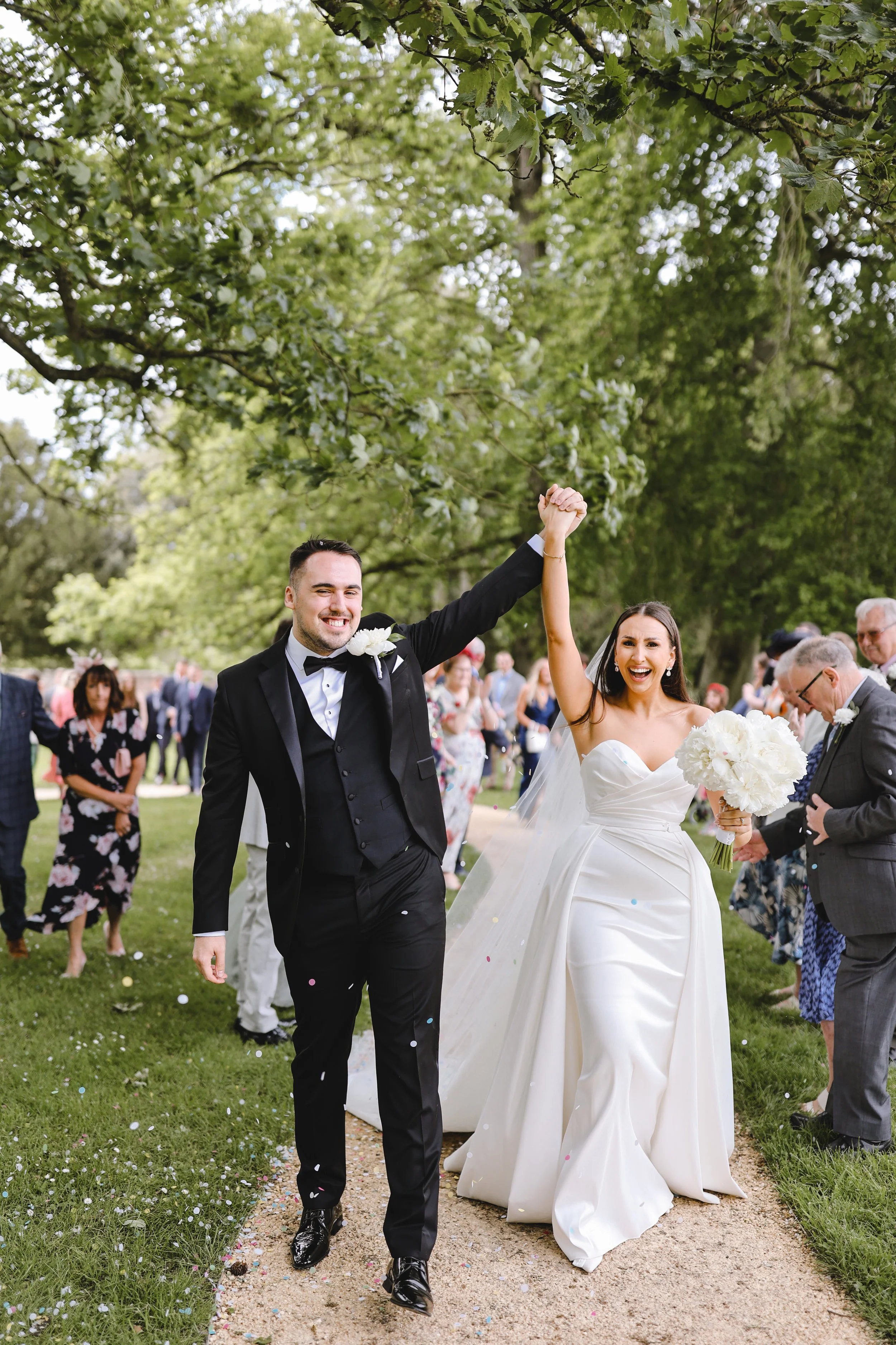 Bride and groom celebrating outdoors, holding hands, surrounded by wedding guests and green trees.
