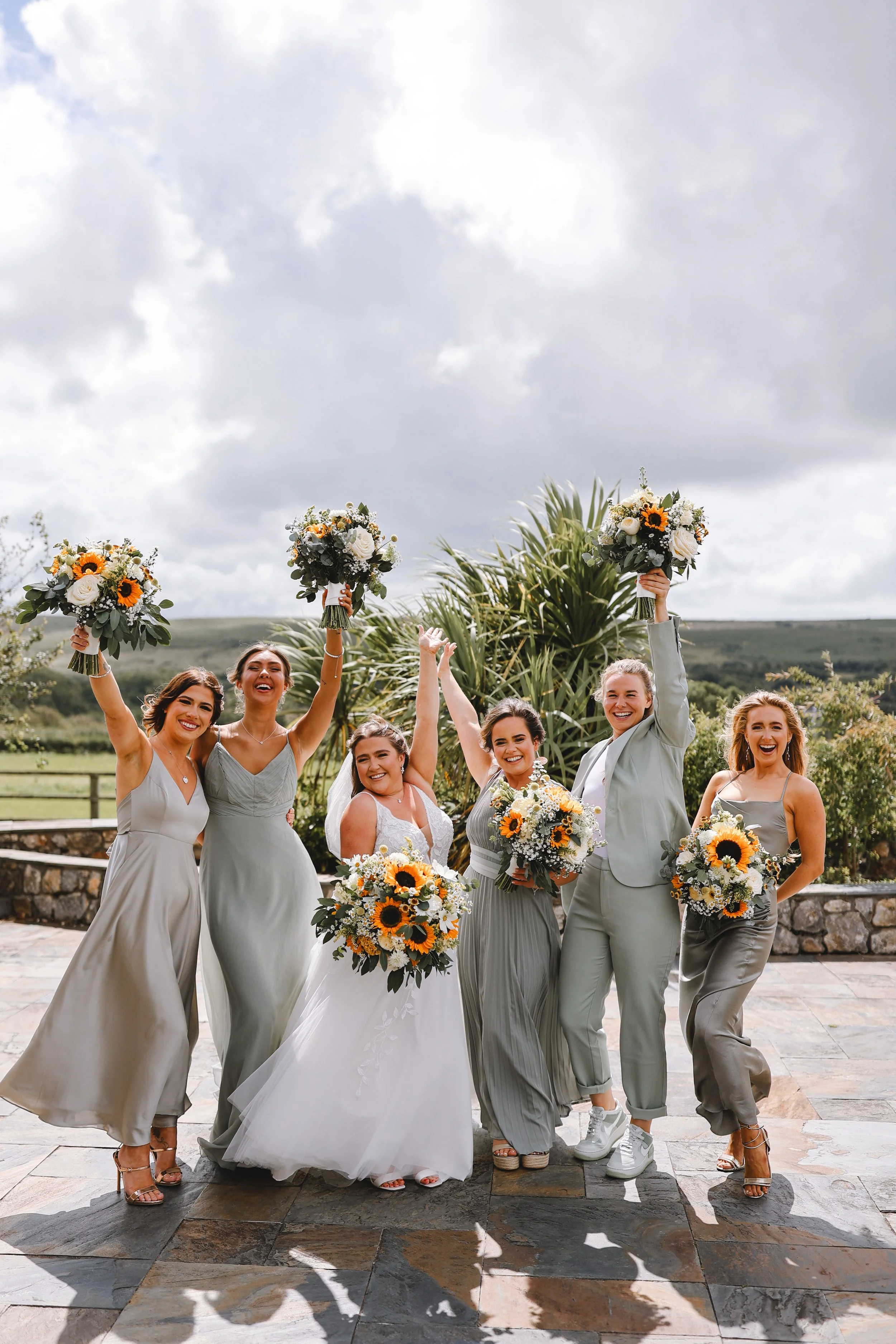 A bride with five bridesmaids poses outdoors in pastel dresses holding bouquets of sunflowers and white flowers. They stand on a patio with a cloudy sky and lush greenery in the background.
