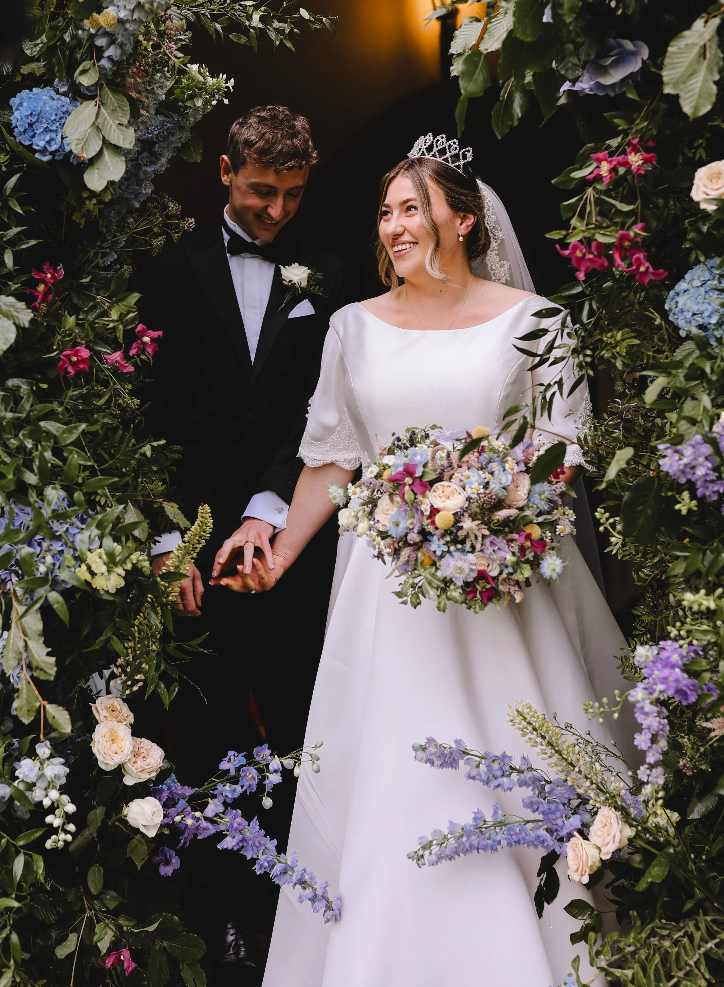 A bride and groom smiling, holding hands, surrounded by colorful flowers. The bride is wearing a white dress and tiara, holding a bouquet. The groom is in a black tuxedo.