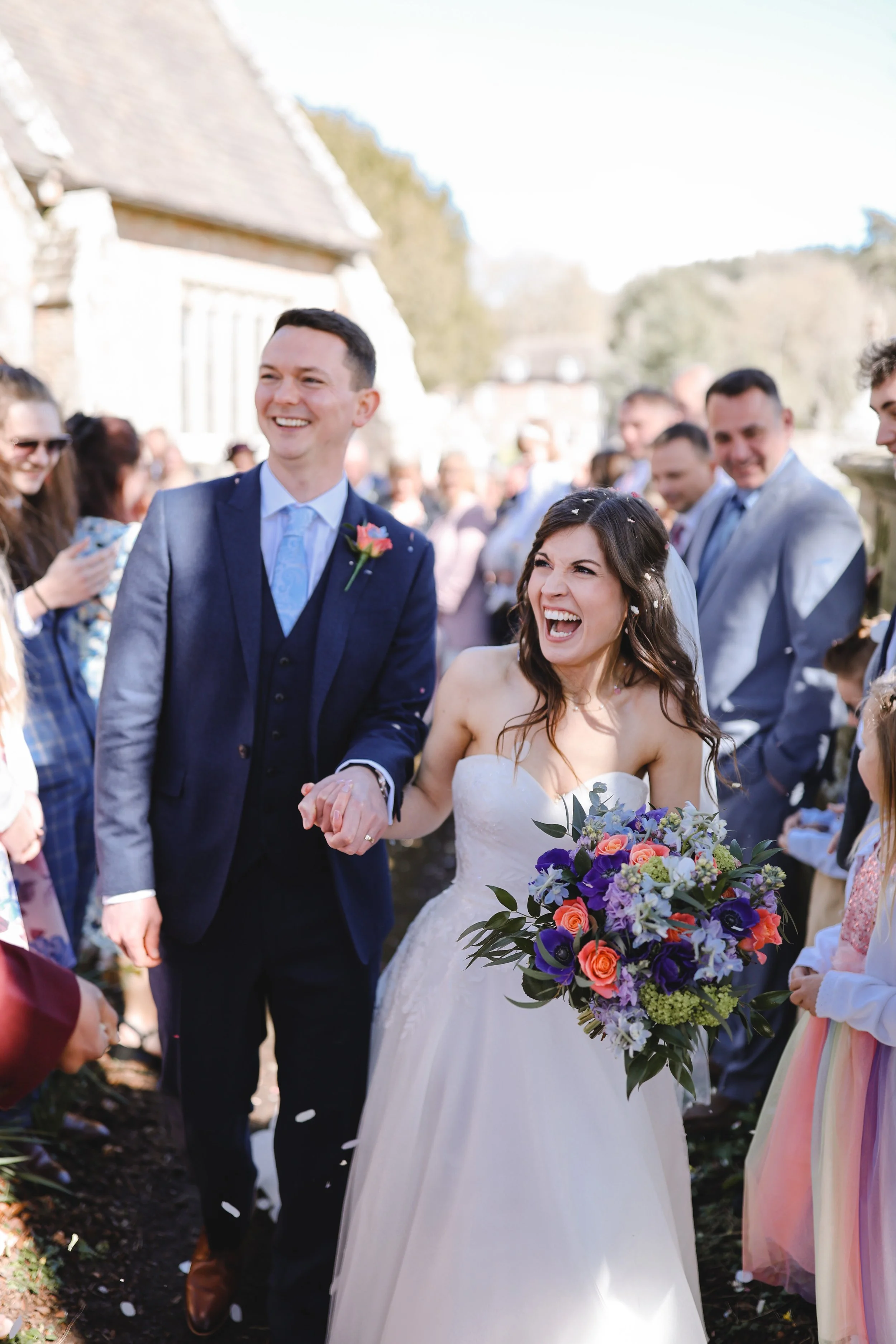 Bride and groom walking joyfully outdoors with a crowd of guests, holding hands. The bride is wearing a white dress, carrying a colorful bouquet, and the groom is in a suit with a boutonnière. People are smiling and celebrating around them.