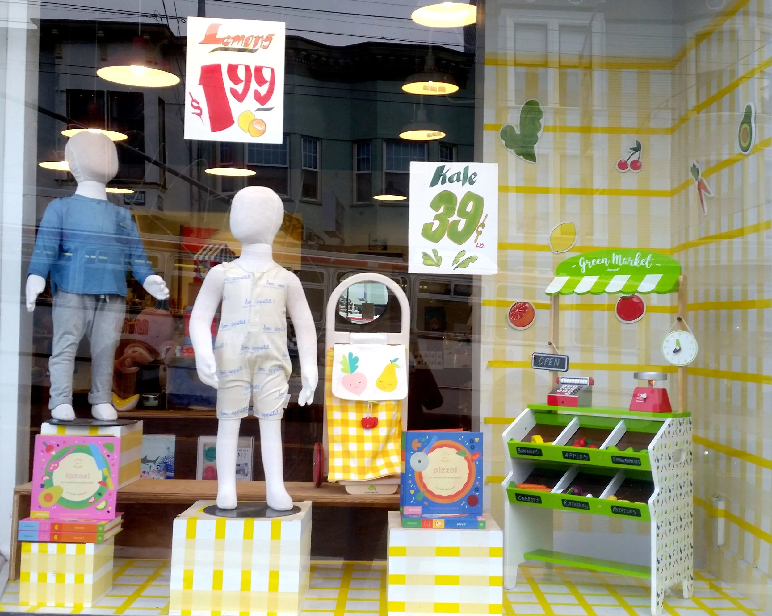Grocery Store inspired window display, featuring yellow plaid on all surfaces, custom-painted signs and fruit/veggie decals and food themed products. Spring/Summer 2018