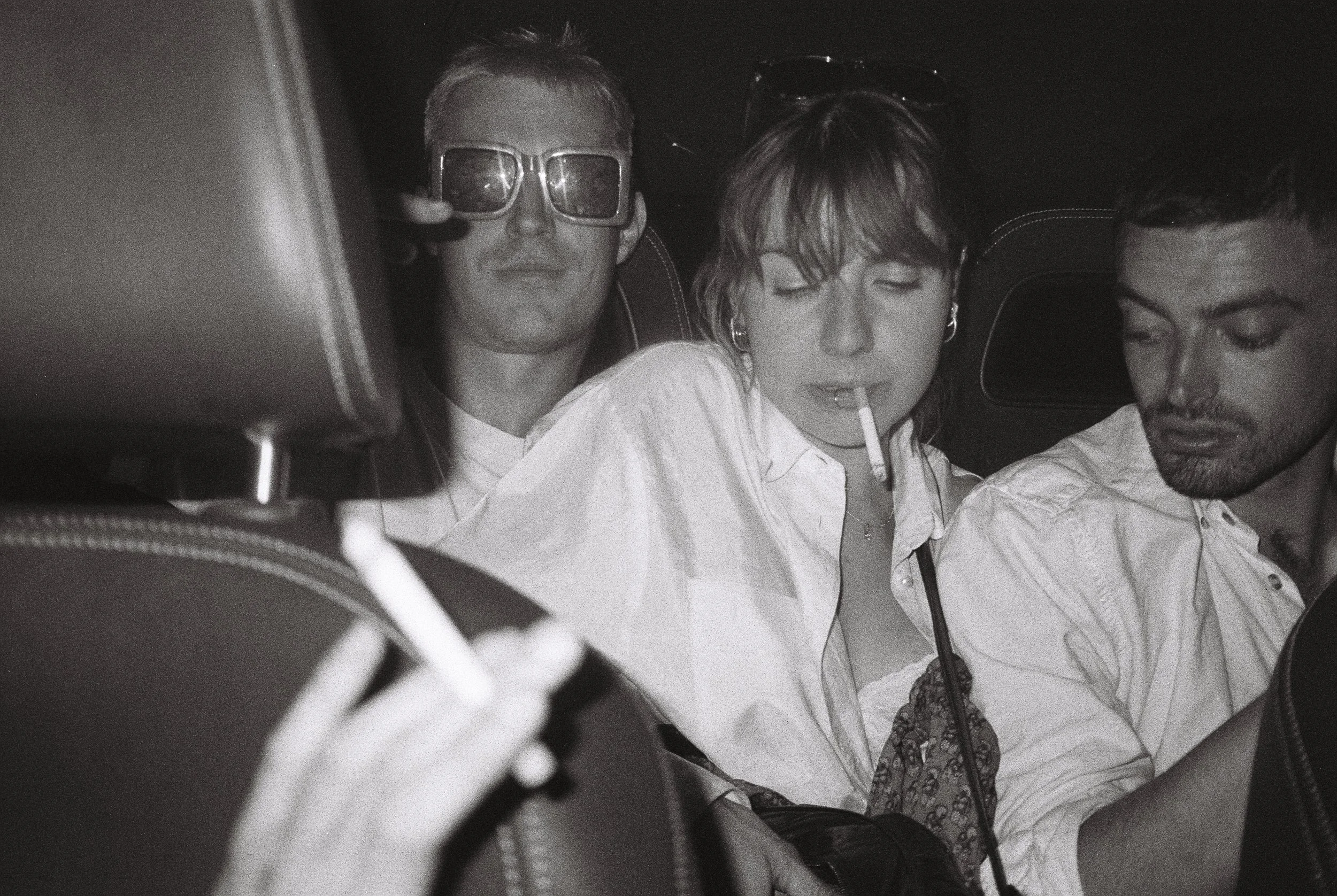 Black and white photo of three young adults taking a selfie, with two men and one woman, inside a car at night.