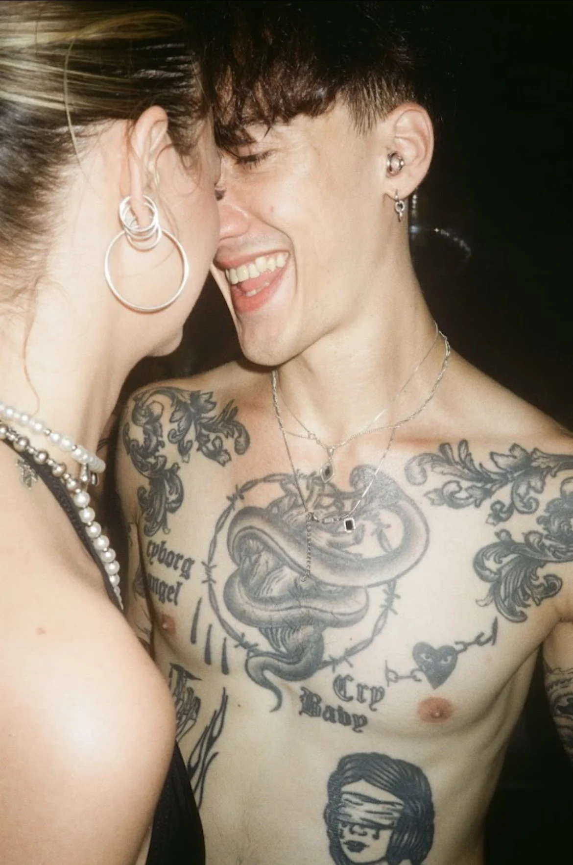 A shirtless man with numerous tattoos on his chest and shoulders, smiling and leaning close to a woman with earrings and a pearl necklace.
