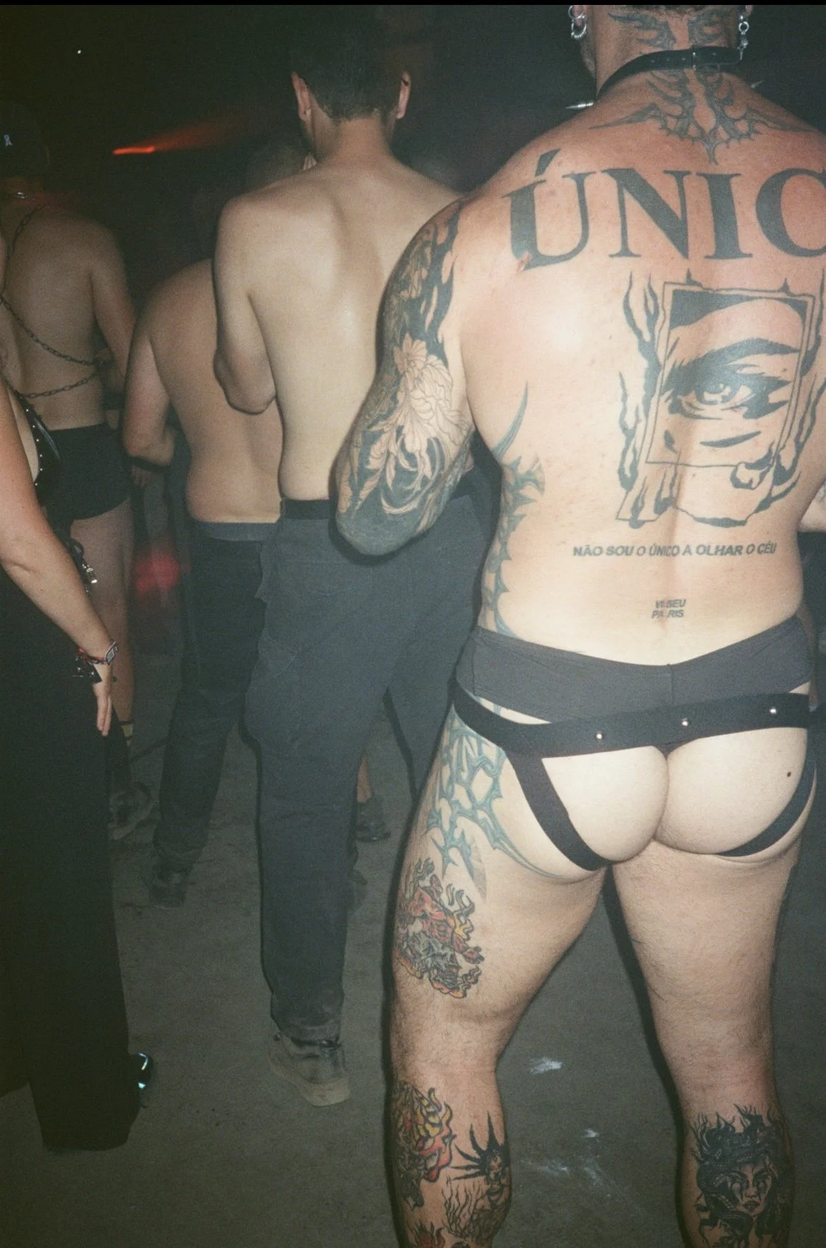 People standing in a line at a nightclub, with one person in the foreground showcasing numerous tattoos and wearing minimal clothing.