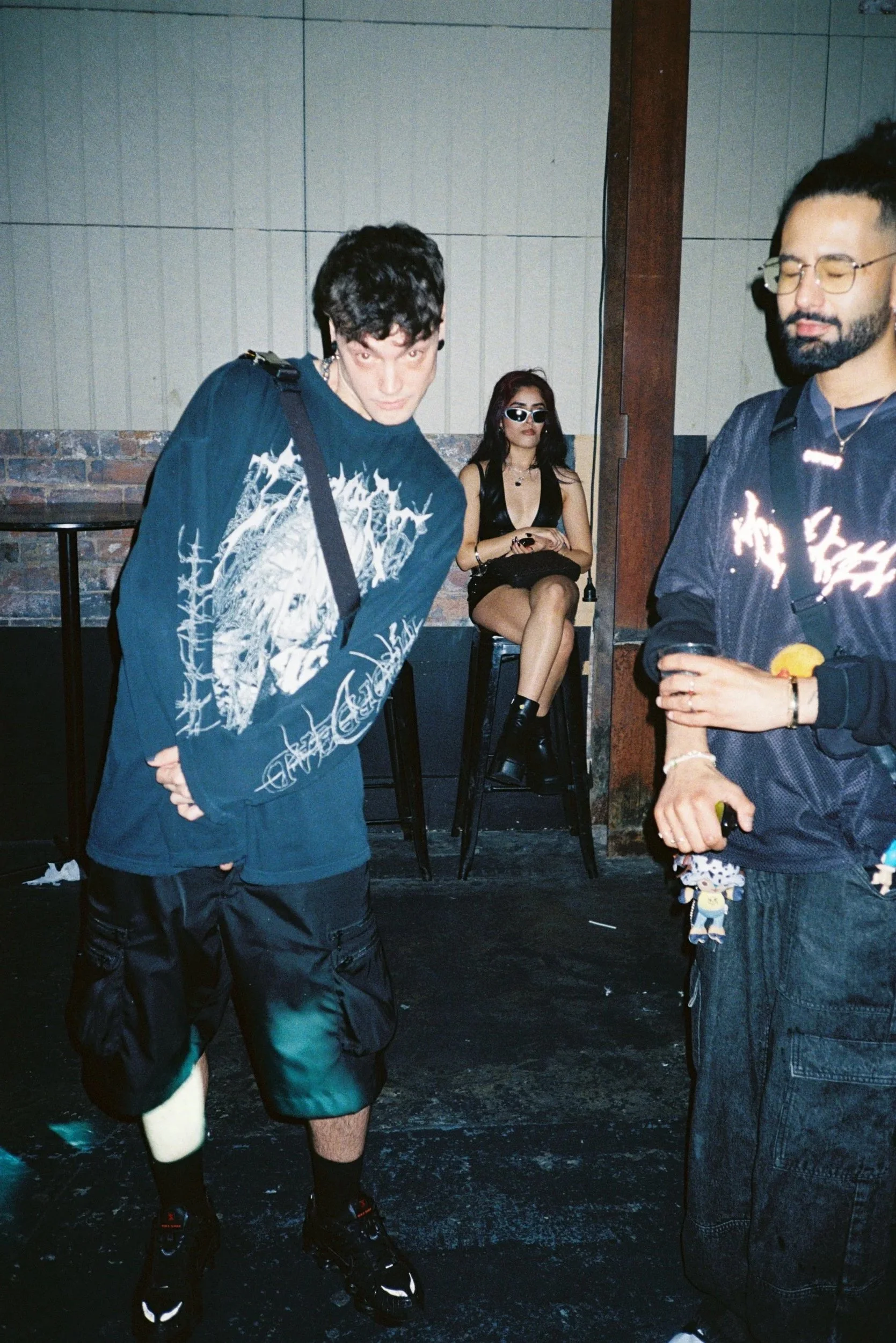 Two men standing and a woman sitting on a barstool in a dimly lit indoor space with brick walls. The woman is wearing sunglasses and a black outfit, while one man has curly hair and a dark graphic shirt, and the other has a beard, glasses, and a dark