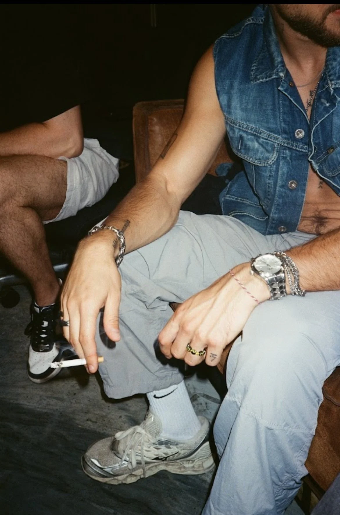 A person wearing a sleeveless denim vest, grey shorts, and a watch, holding a cigarette, sitting on a brown chair with another person partially visible nearby.