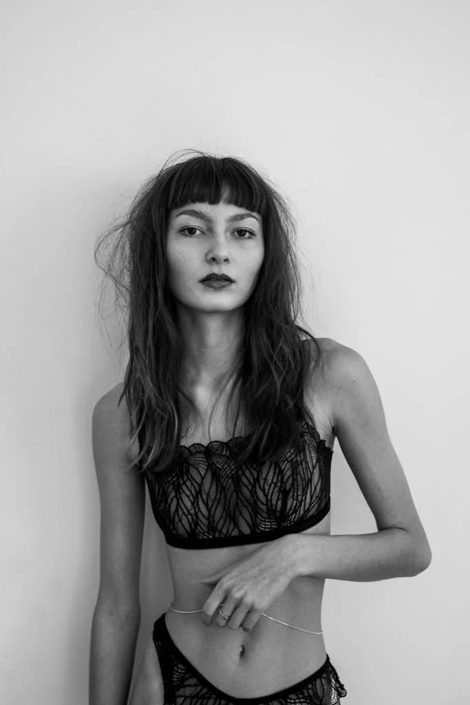 Black and white photograph of a woman with wavy hair and bangs, wearing lace lingerie, standing against a plain wall.