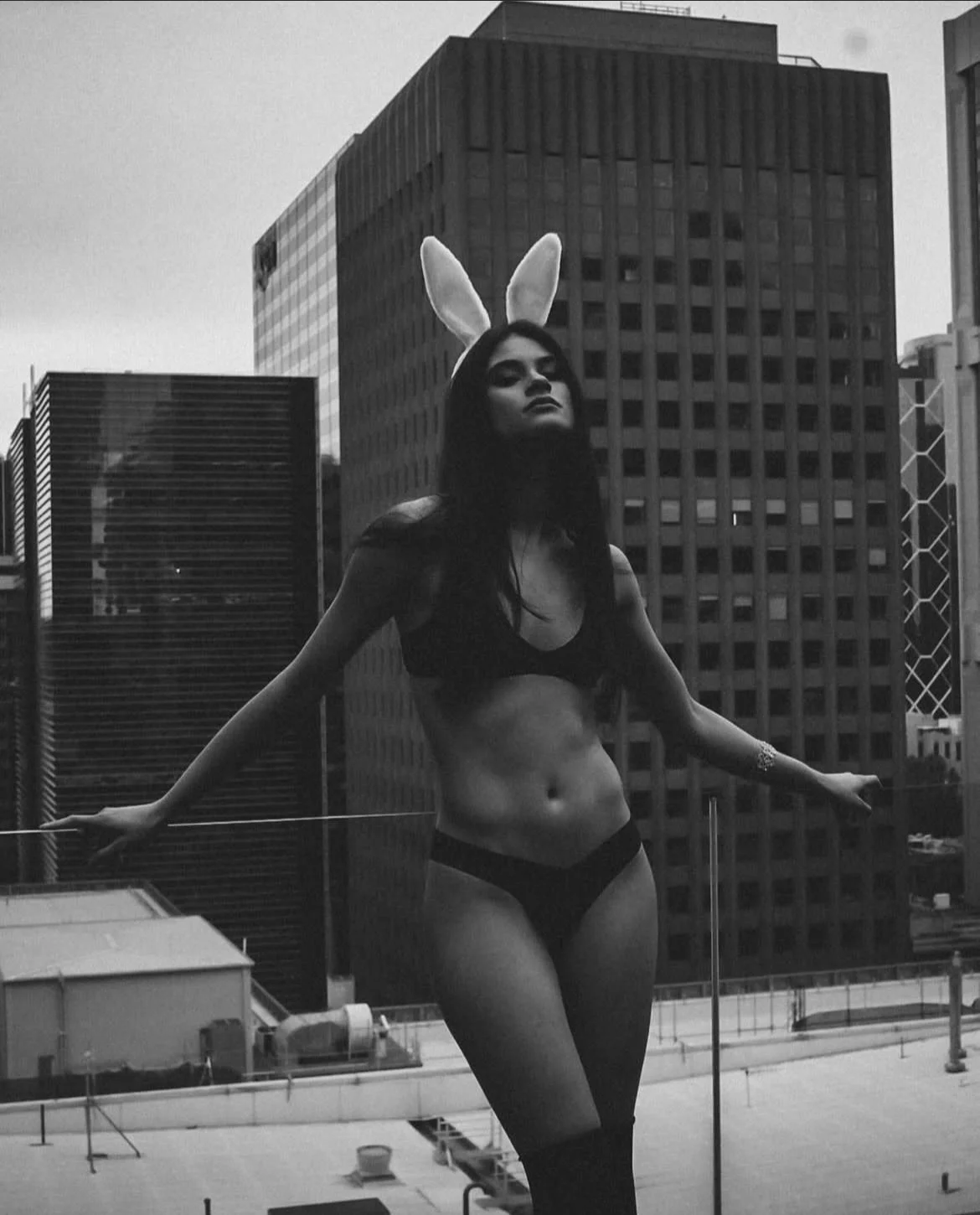 A woman with bunny ears headband standing on a rooftop in front of skyscrapers, wearing a black crop top and underwear, black stockings, with her arms outstretched.