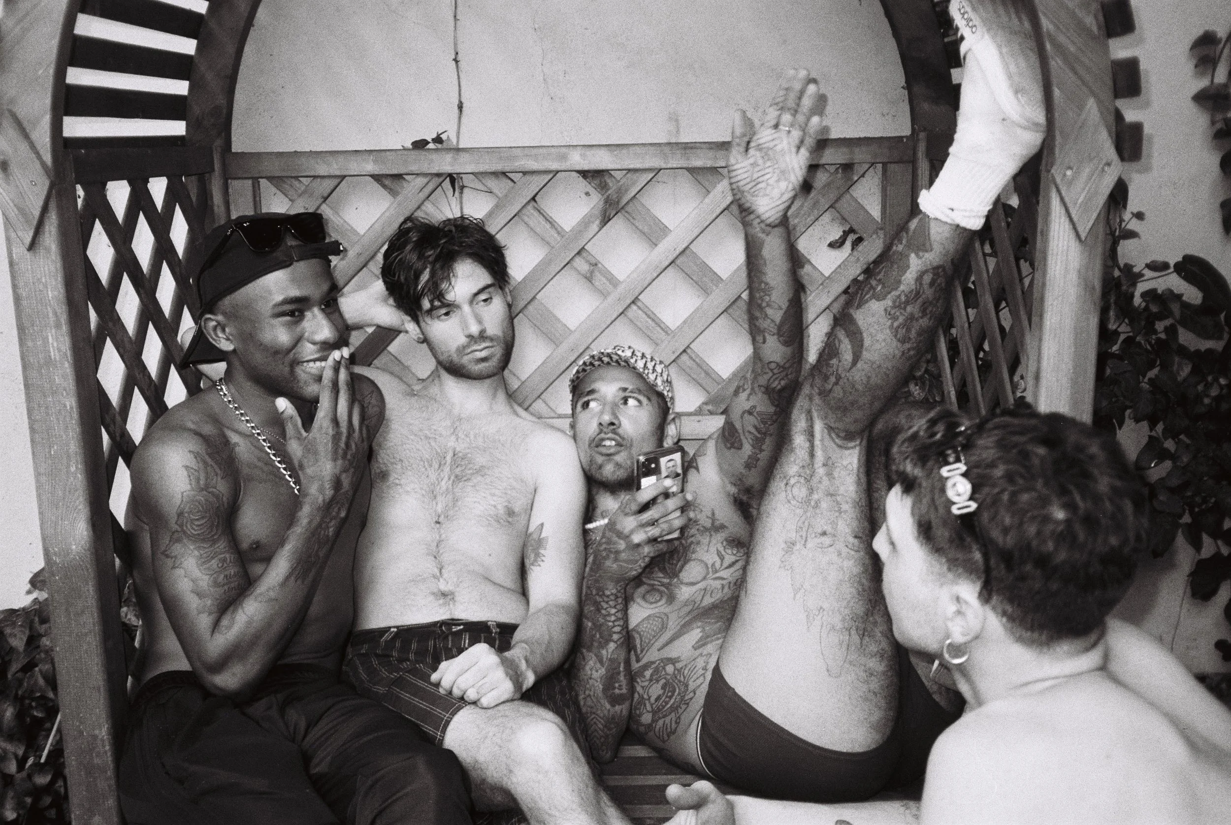 Five people sitting on a wooden bench in a garden, four shirtless and one with his back to the camera. They are engaging in a conversation and posing for a photo, with one person holding a smartphone. The scene is casual and relaxed.