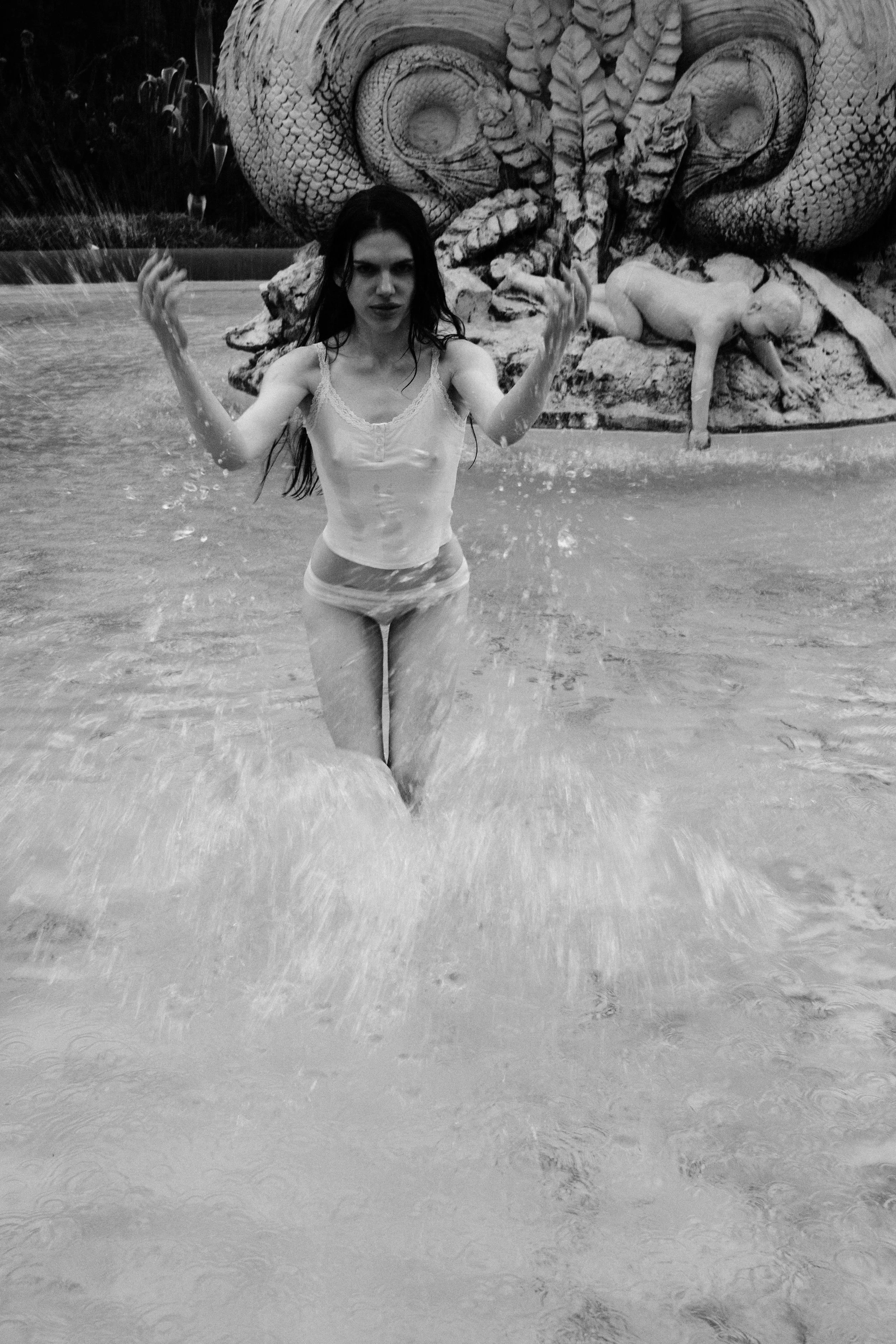 A woman in a swimsuit standing in a pool with her arms raised, in front of a large sculpture of a snake's head and a dog figure.