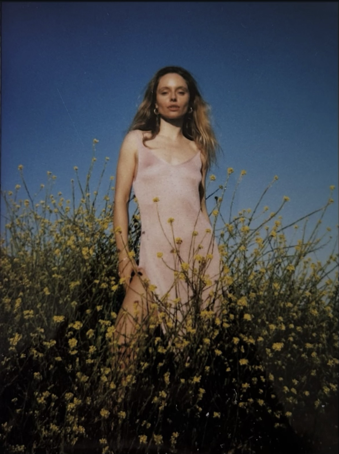 A woman with long hair wearing a light pink dress standing in a field of yellow flowers under a clear blue sky.