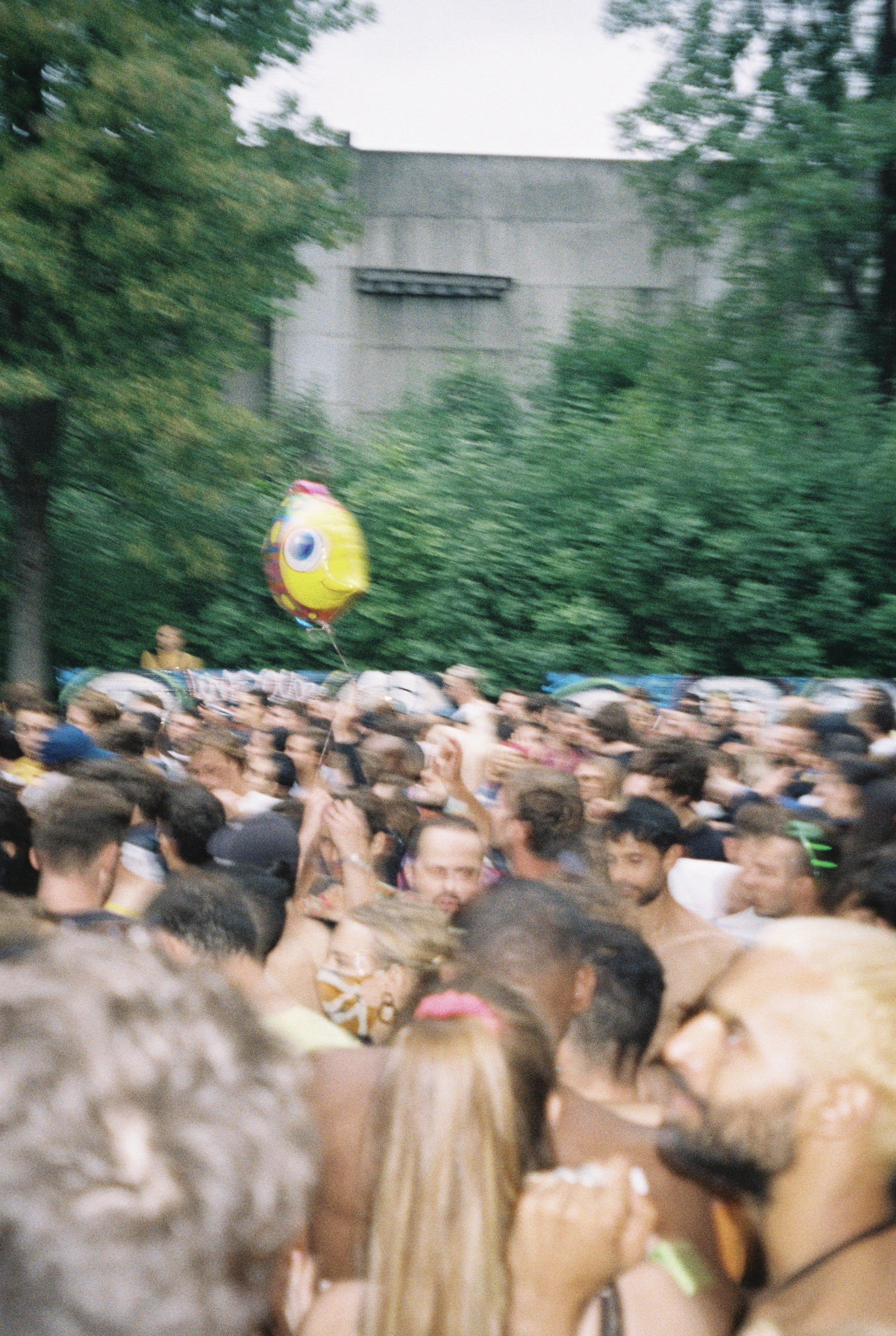 Crowd of people at an outdoor event, some are wearing swimsuits and drinking, with a colorful fish-shaped balloon floating above; trees and an industrial building are in the background.