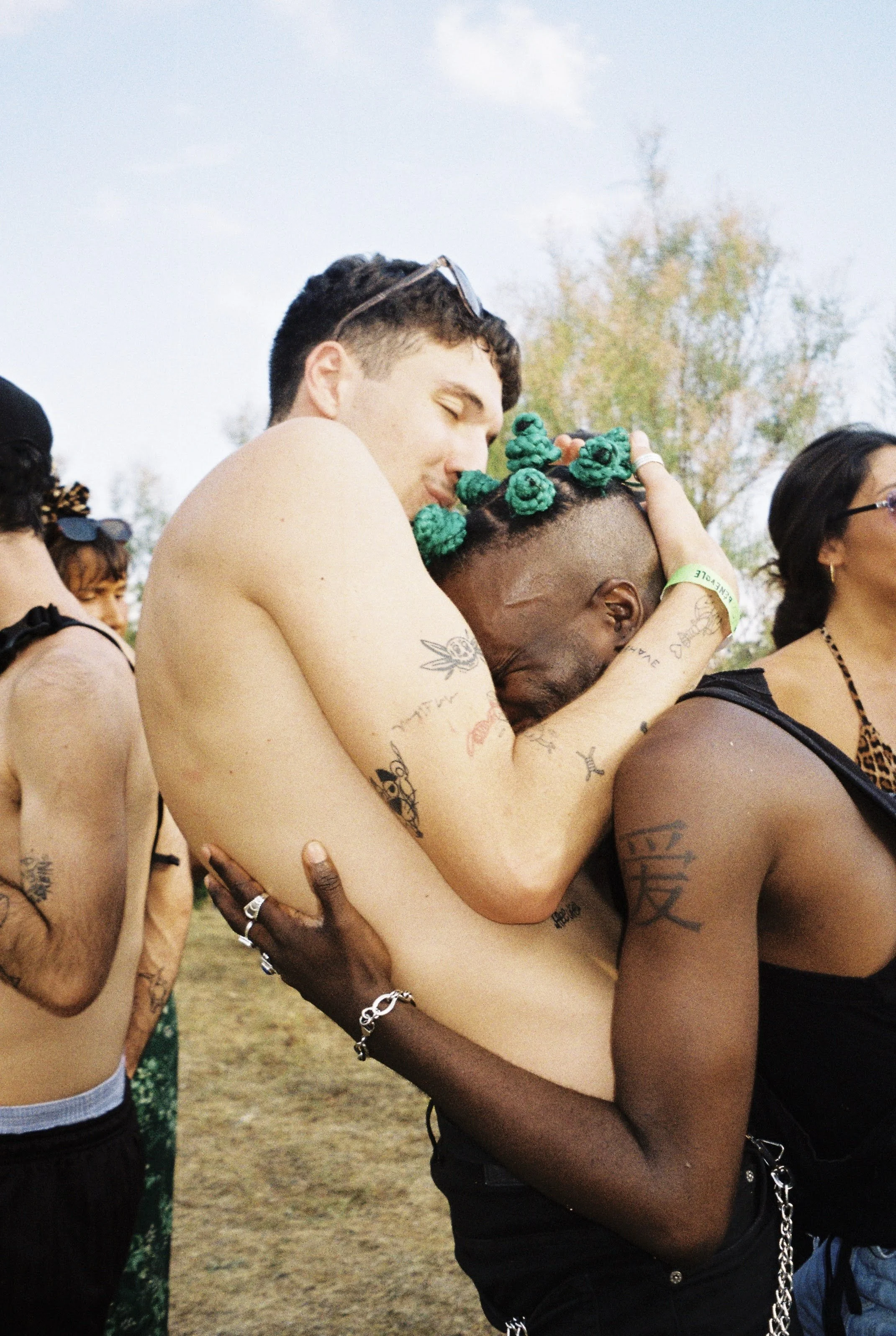 Two men embracing at an outdoor event, one with multiple green hair accessories, tattoos, and wearing a bracelet, while the other is shirtless with sunglasses on his head, and other people are visible in the background.