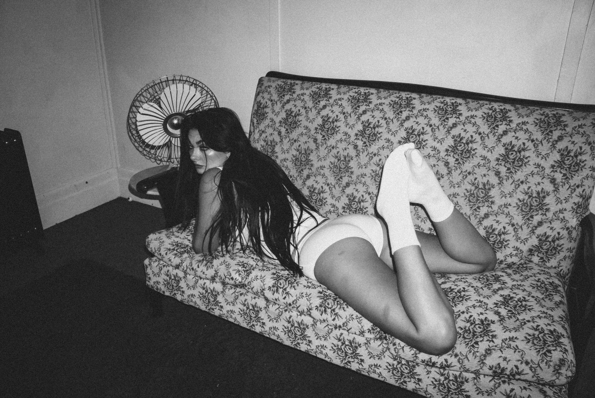 A woman with long dark hair lying on a floral-patterned couch, wearing white underwear and knee-high socks, in a black and white photograph.