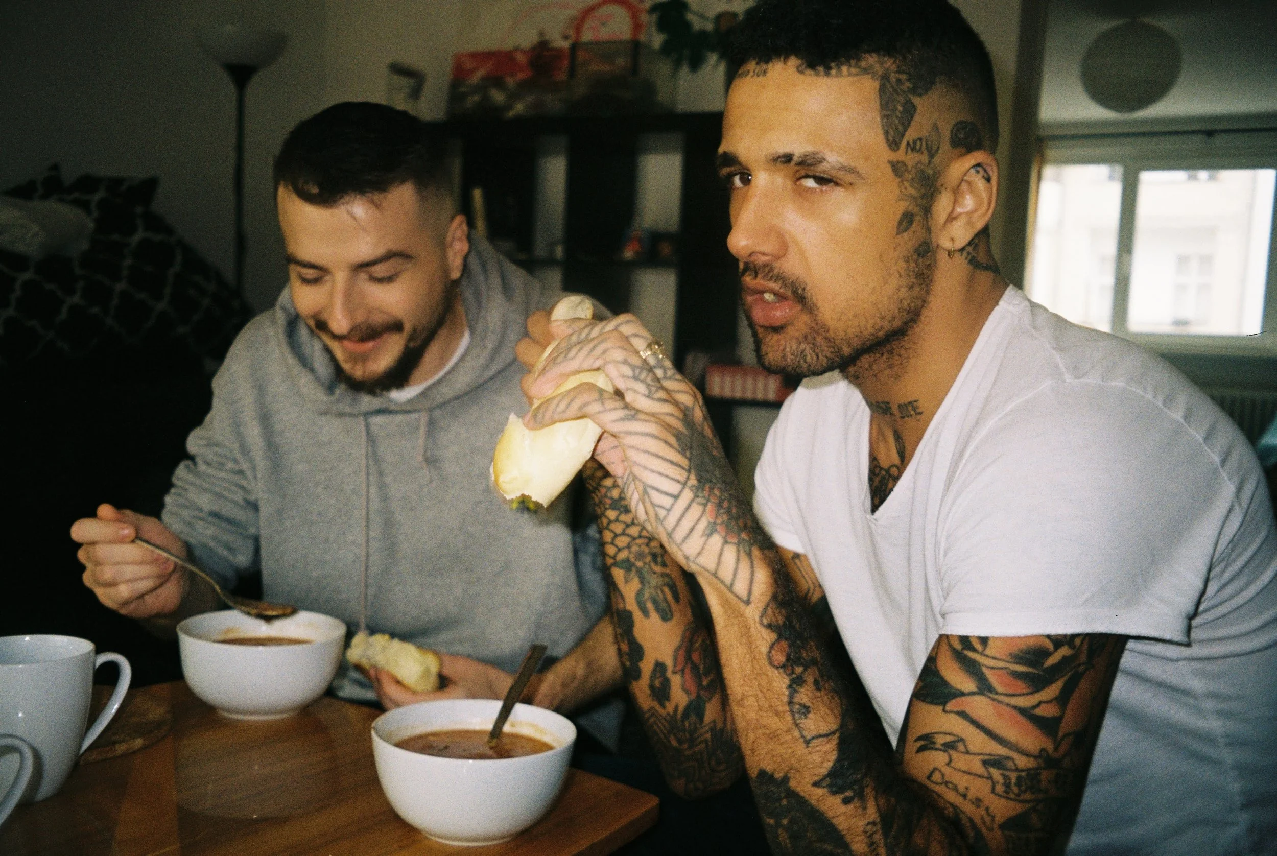 Two men sitting at a table, eating soup from white bowls. One man is smiling and holding bread, the other man is wearing a white T-shirt, has tattoos, and is holding bread while looking at the camera.