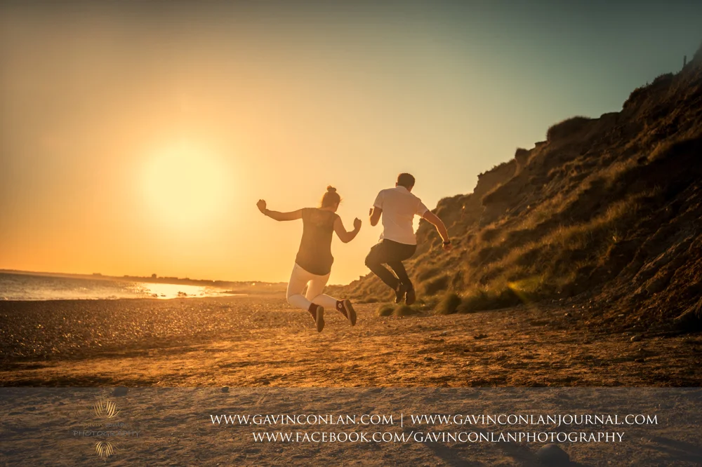 fun portrait of Victoria and James jumping in the air on the beach of Hengistbury Head at sunset. Engagement Session in Bournemouth, Dorset by gavin conlan photography Ltd