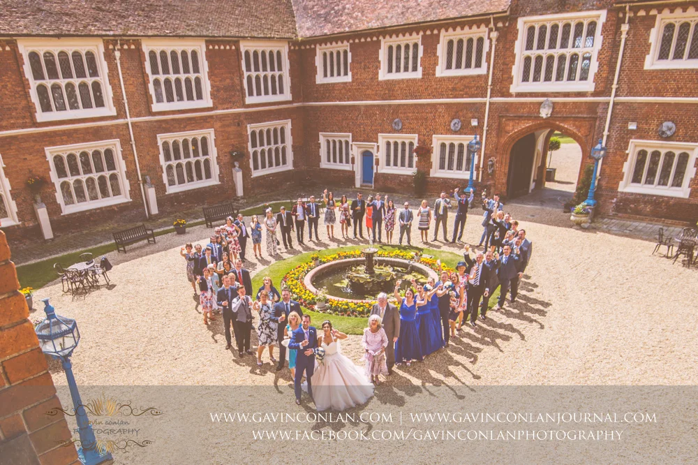 creative group photograph of the bride and groom with all their guests standing around the fountain in a heart shaped in the inner courtyard of Gosfield Hall. Wedding photography at Gosfield Hall by Essex wedding photographer gavin conlan photography Ltd