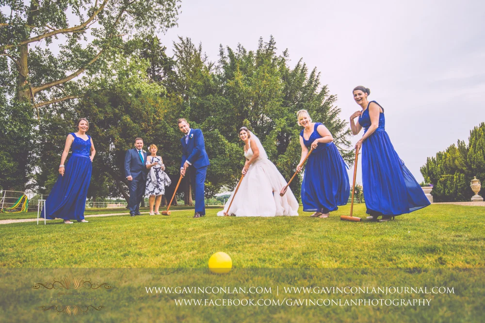 fun portrait of the bride, groom and the bridesmaids playing croquet on the lawn outside the main entrance of Gosfield Hall with the grooms mother and step dad looking on in the background. Wedding photography at Gosfield Hall by Essex wedding photographer gavin conlan photography Ltd
