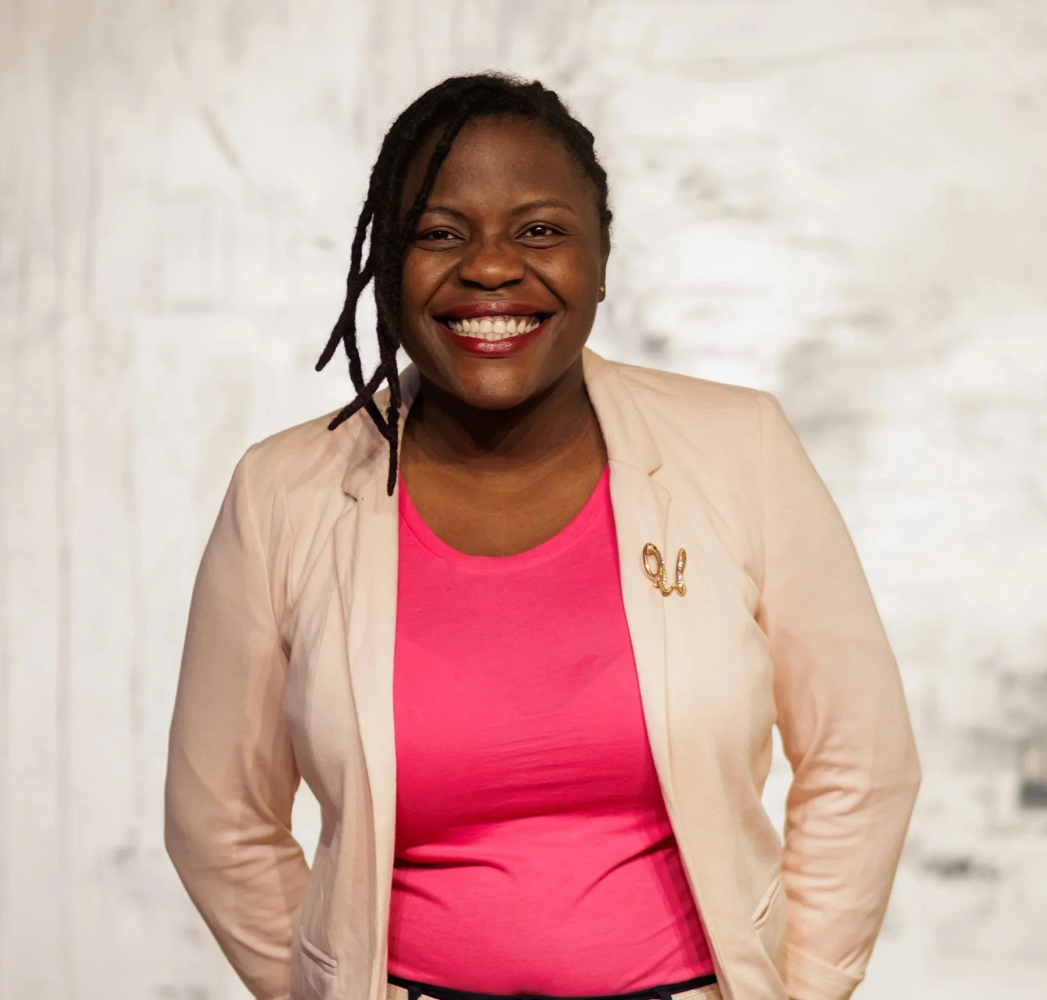 Gemini said A smiling woman with dark locs wearing a bright pink top and a light beige blazer.