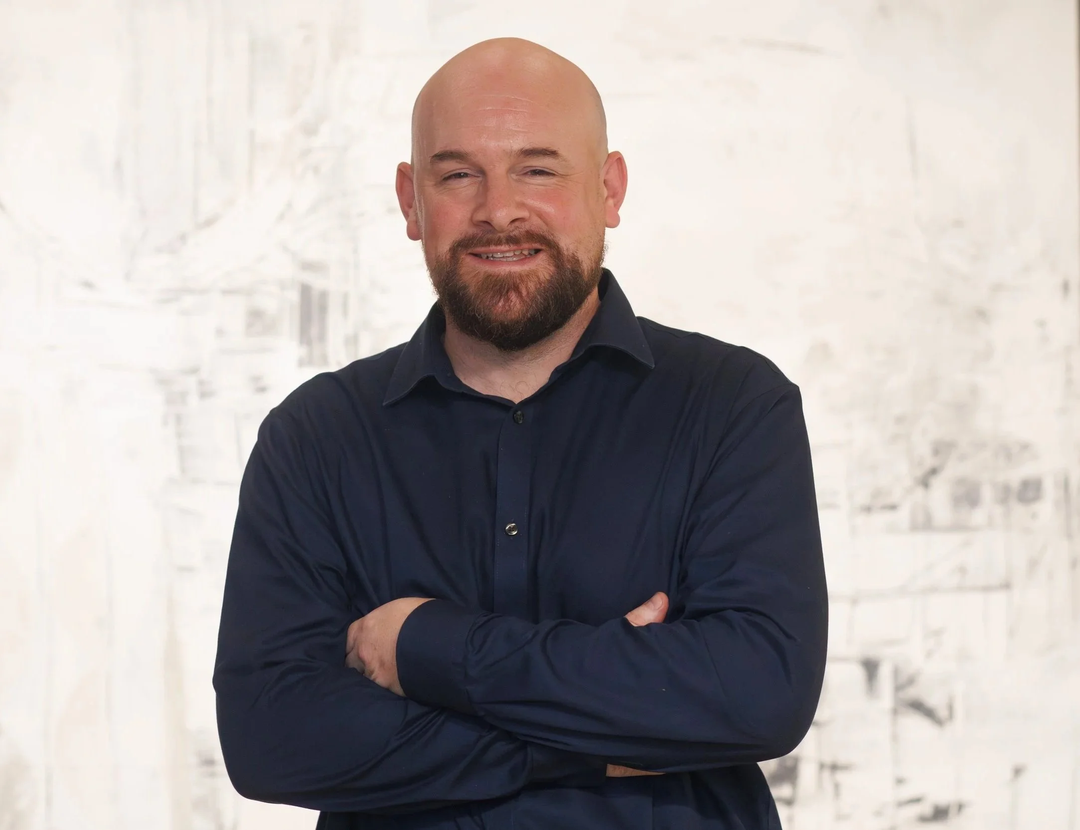A medium-close up photograph of a bald, smiling man with a beard wearing a dark blue button-up shirt with his arms crossed.