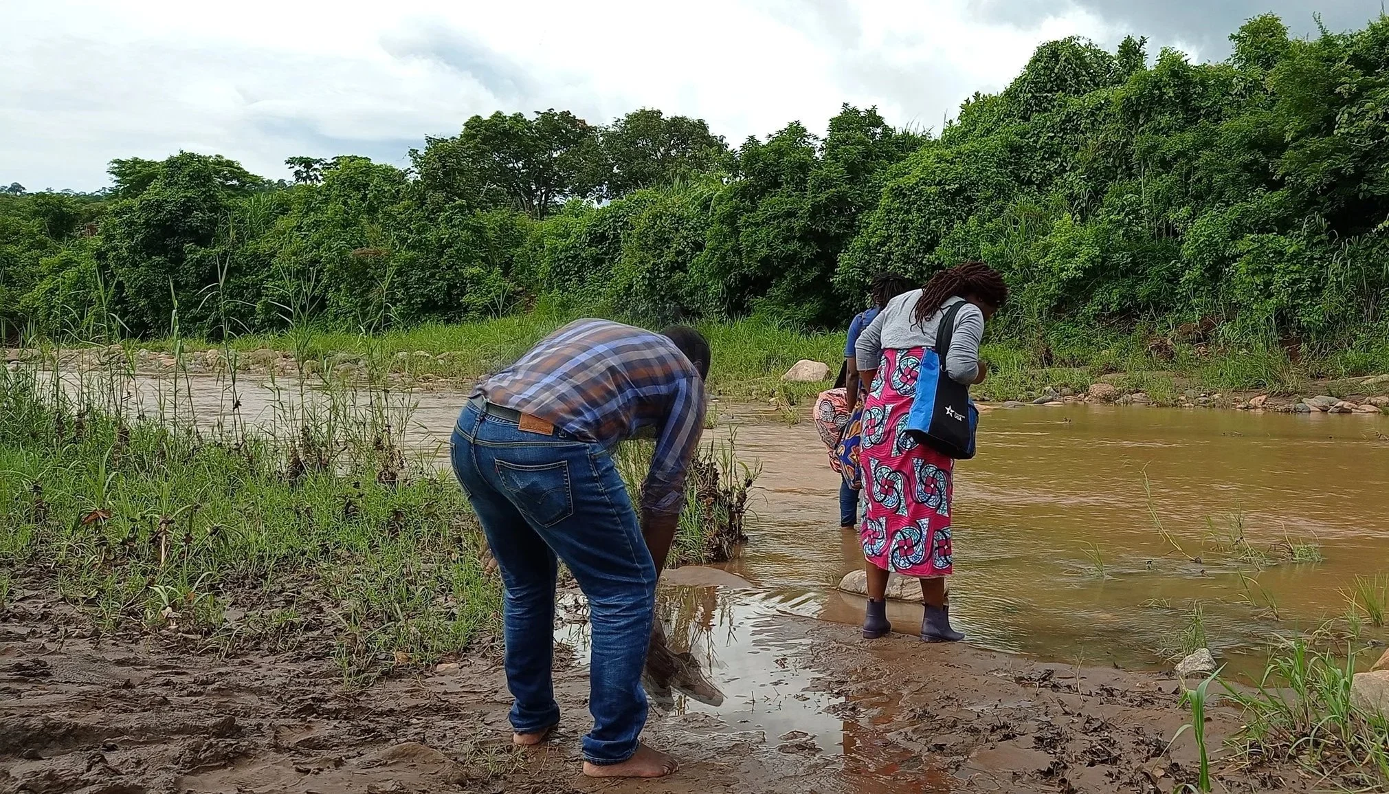 A candid shot of three people at a muddy riverbank under an overcast sky. One man in a plaid shirt bends over the shore, while another person stands ankle-deep in the turbid water. Dense green foliage lines the far bank.
