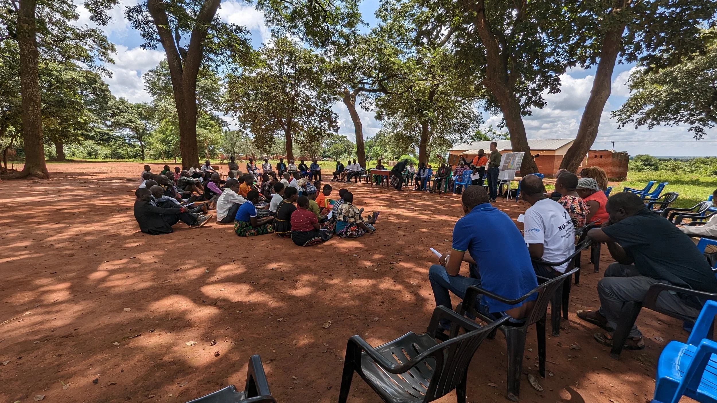 A large group of community members gathered outdoors under the shade of tall trees for a meeting. Many are seated on the ground in the foreground, while others sit on chairs in a semi-circle facing a small table where a facilitator is standing.