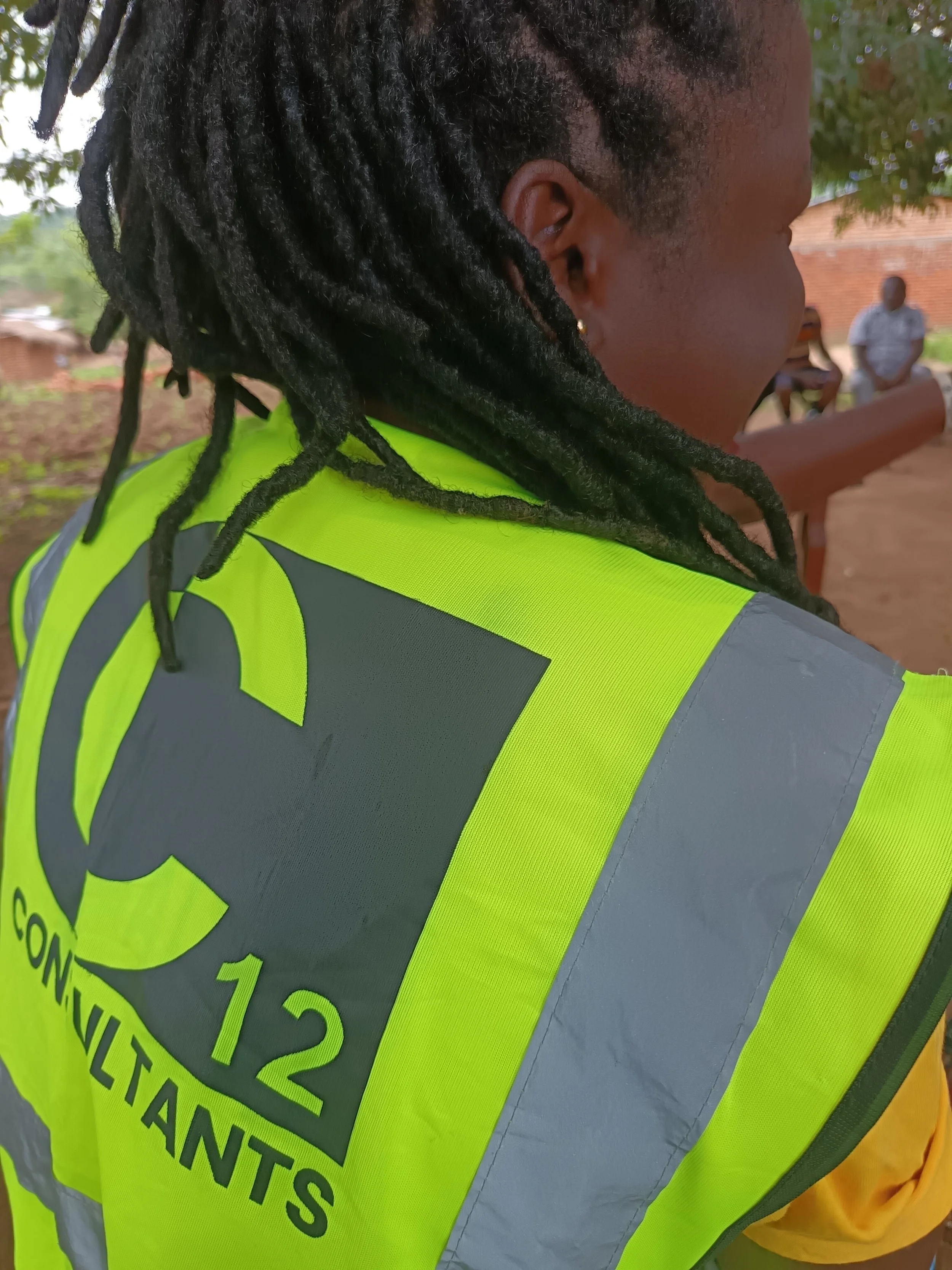 A close-up, over-the-shoulder shot of a team member wearing a high-visibility neon yellow safety vest with reflective gray stripes. The back of the vest prominently features the "C12 Consultants" logo in dark green.