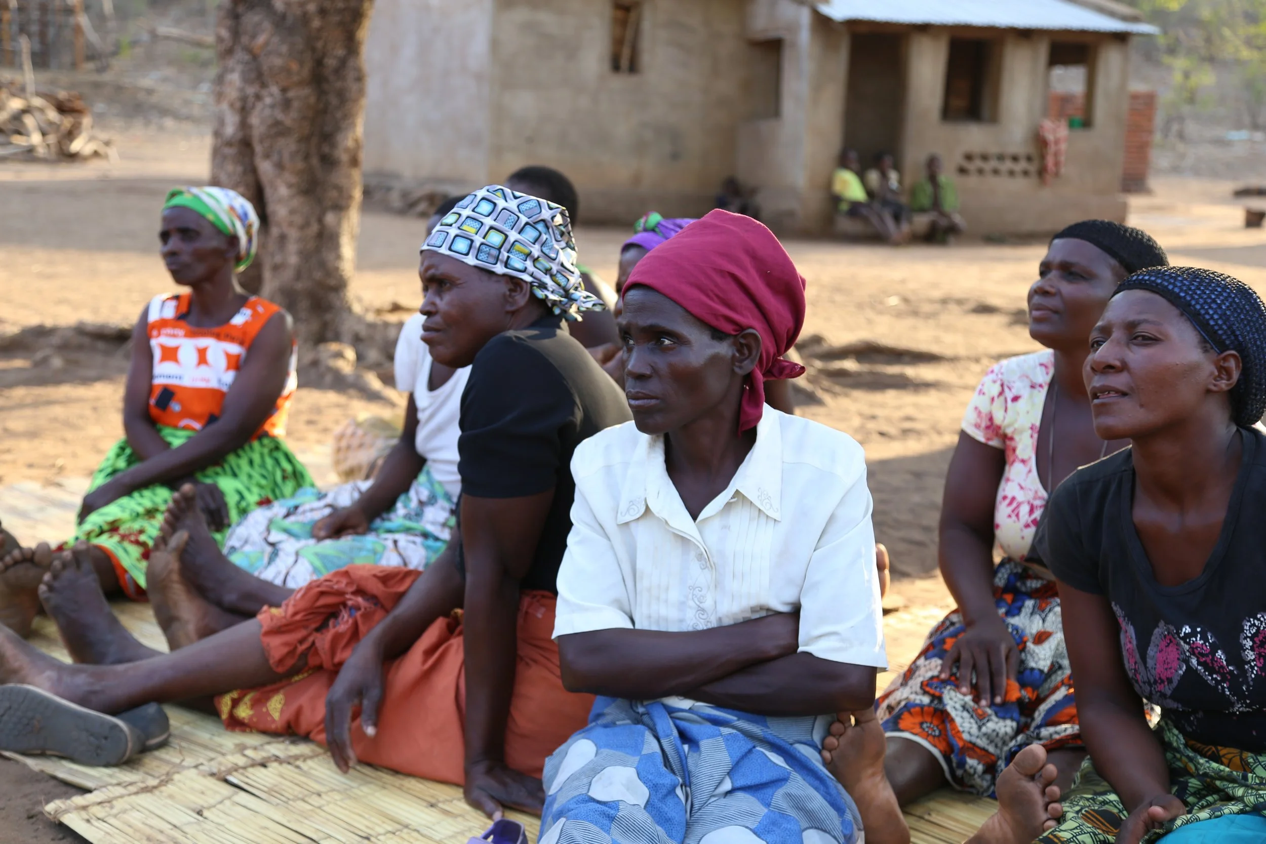 A group of women sitting together on woven mats during an outdoor community meeting. They are dressed in colorful traditional wraps and headscarves, looking attentively toward a speaker off-camera.