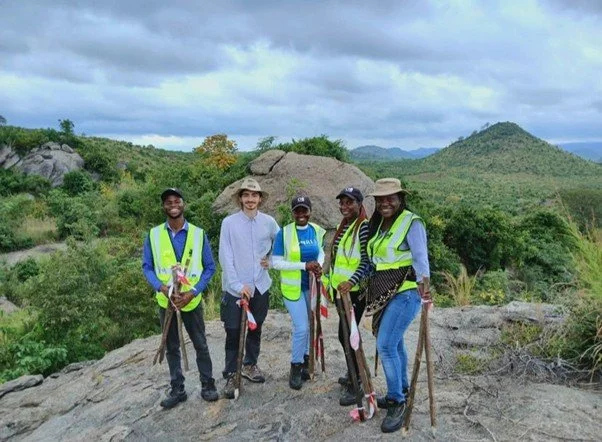 A group of five people posing for a photo on a rocky hilltop. Four team members wear bright neon-yellow safety vests and hats, while several hold wooden walking sticks with red and white markers.