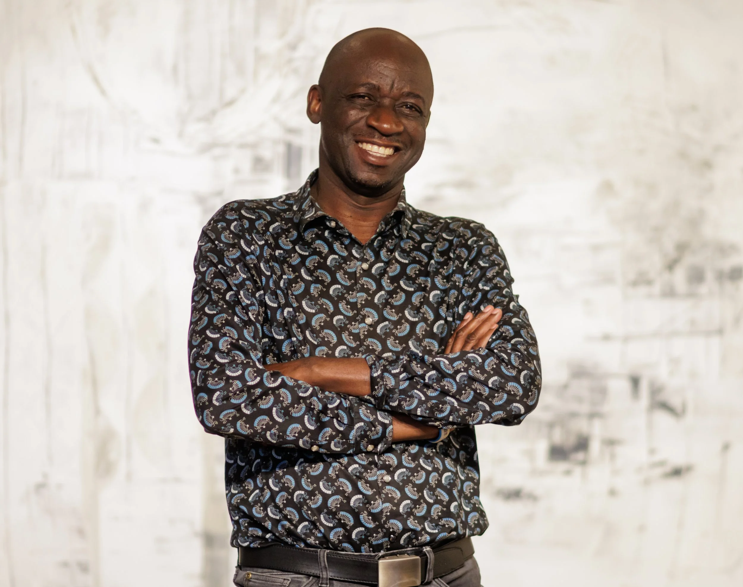 Gemini said A smiling man with a shaved head and crossed arms, wearing a black button-down shirt with a blue and white patterned print.