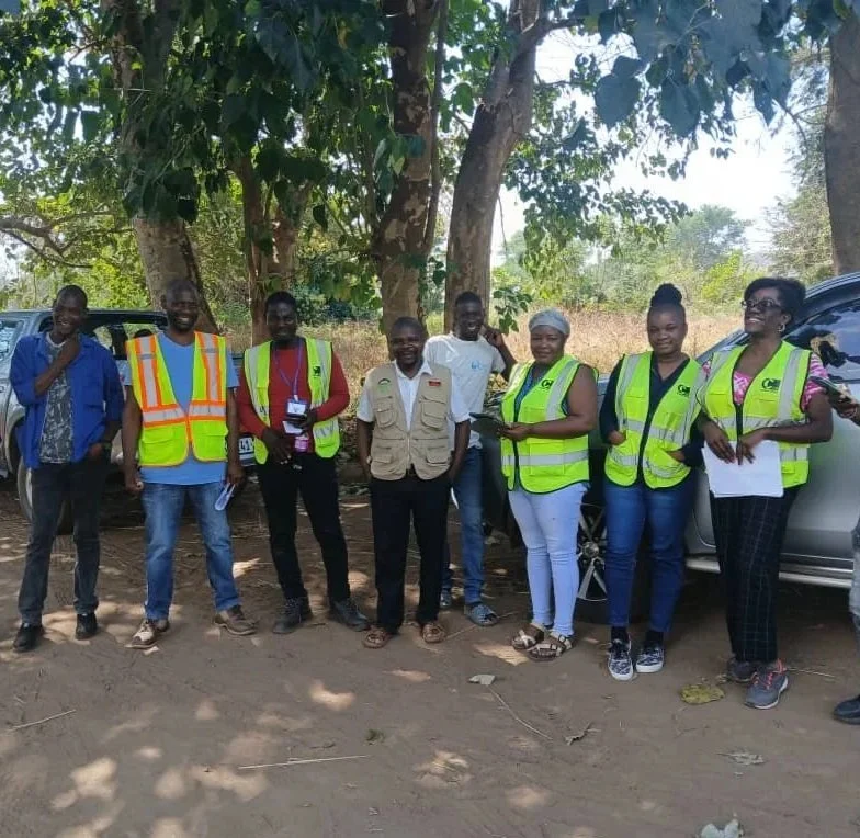 A group of seven people stand together outdoors, posing for a photo under the shade of trees. Most are wearing high-visibility safety vests or utility gear, and two pickup trucks are partially visible behind them.