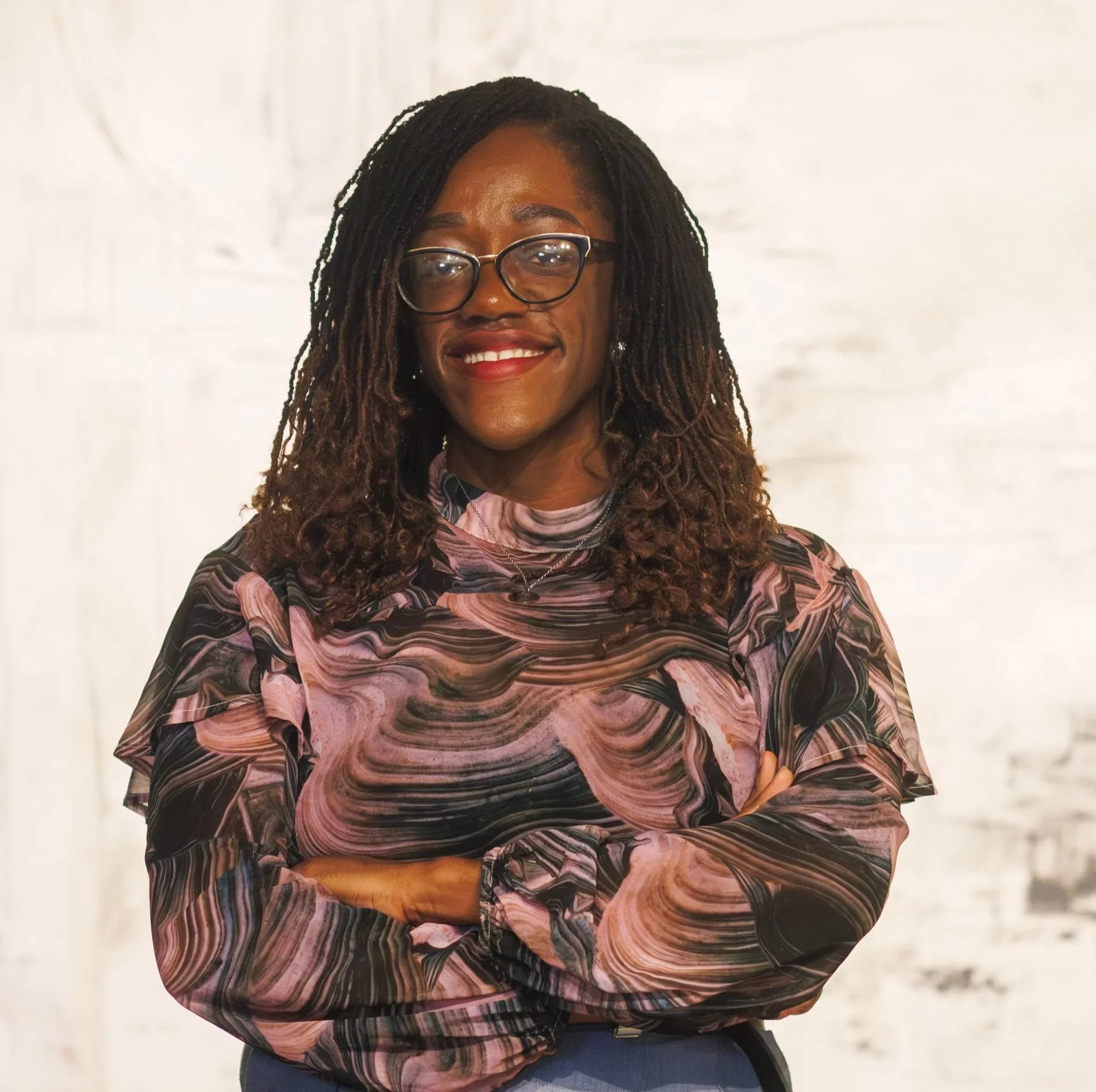 A smiling woman with long sisterlocks and glasses, wearing a patterned pink and black blouse with her arms crossed.