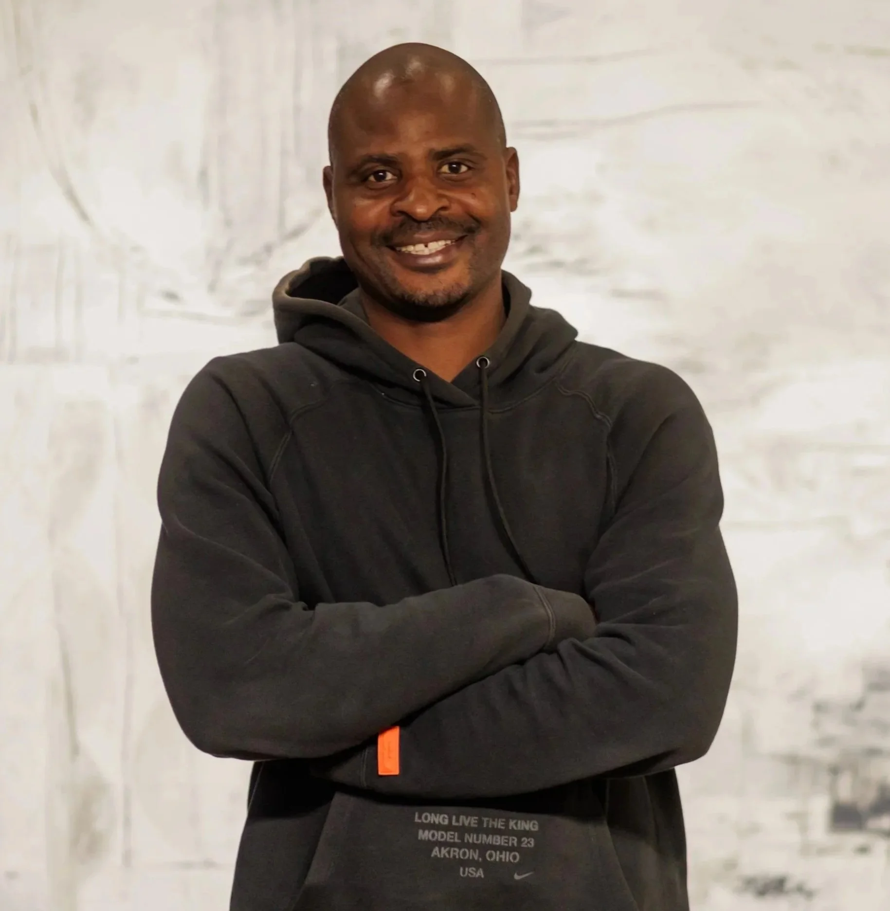 A smiling man with a shaved head and crossed arms, wearing a black hoodie with subtle text on the front pocket.