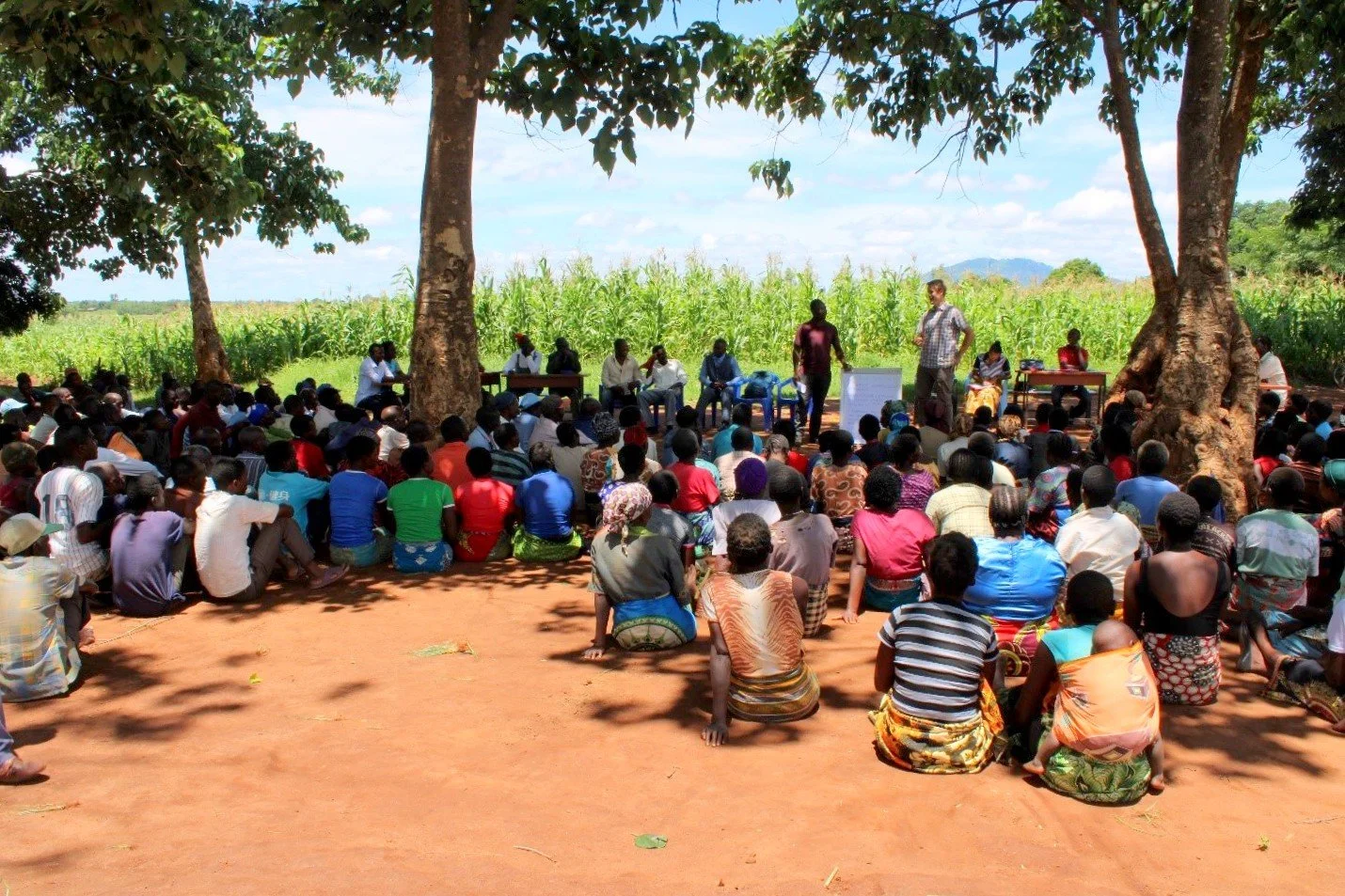 A large group of people sitting on the ground in a semi-circle under the shade of large trees, listening to two men presenting with a whiteboard. A lush green field of crops and distant mountains are visible in the background under a bright sky.