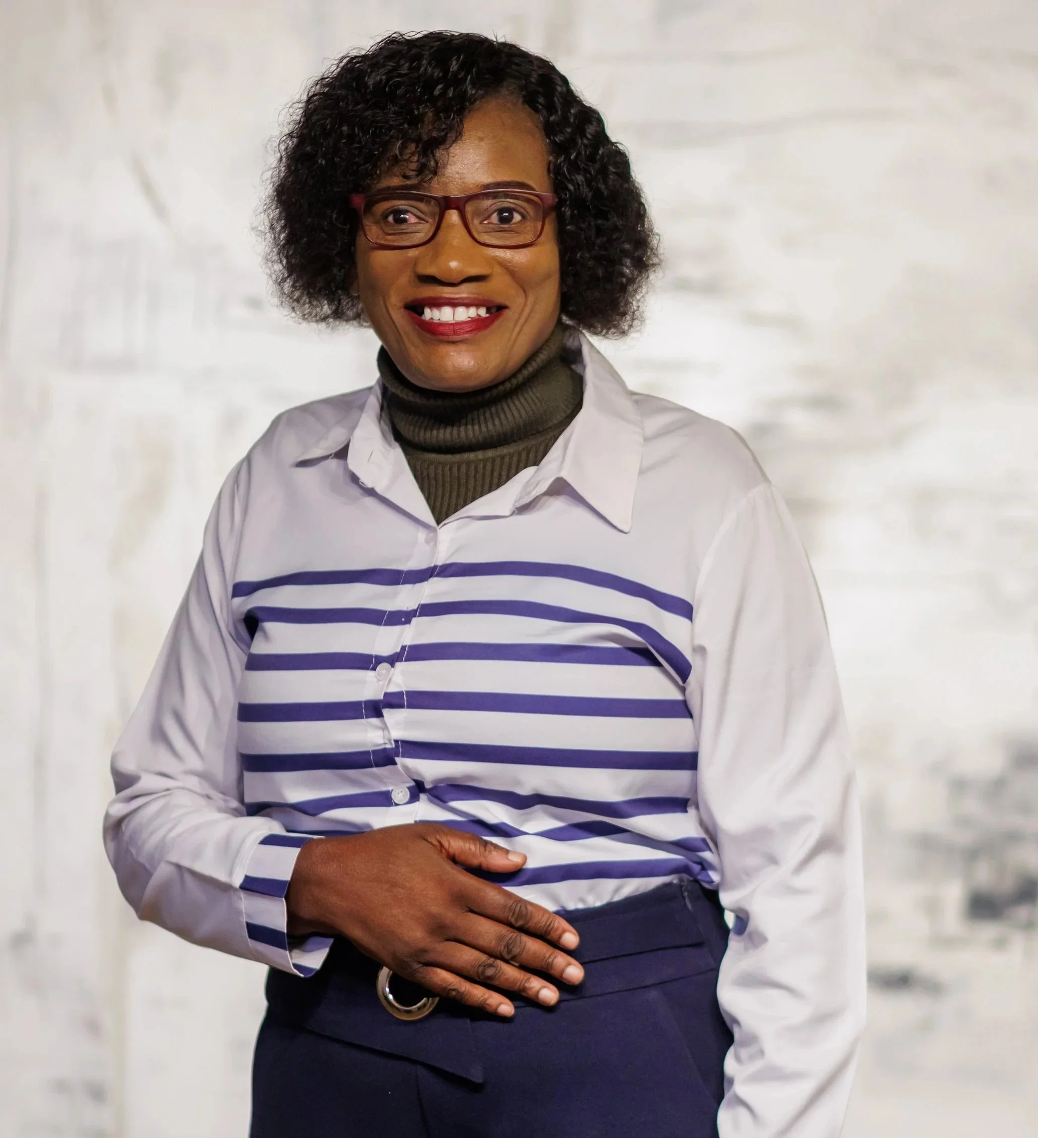 Gemini said A smiling woman with short, curly dark hair and glasses, wearing a white and blue striped shirt over a dark turtleneck and navy trousers.