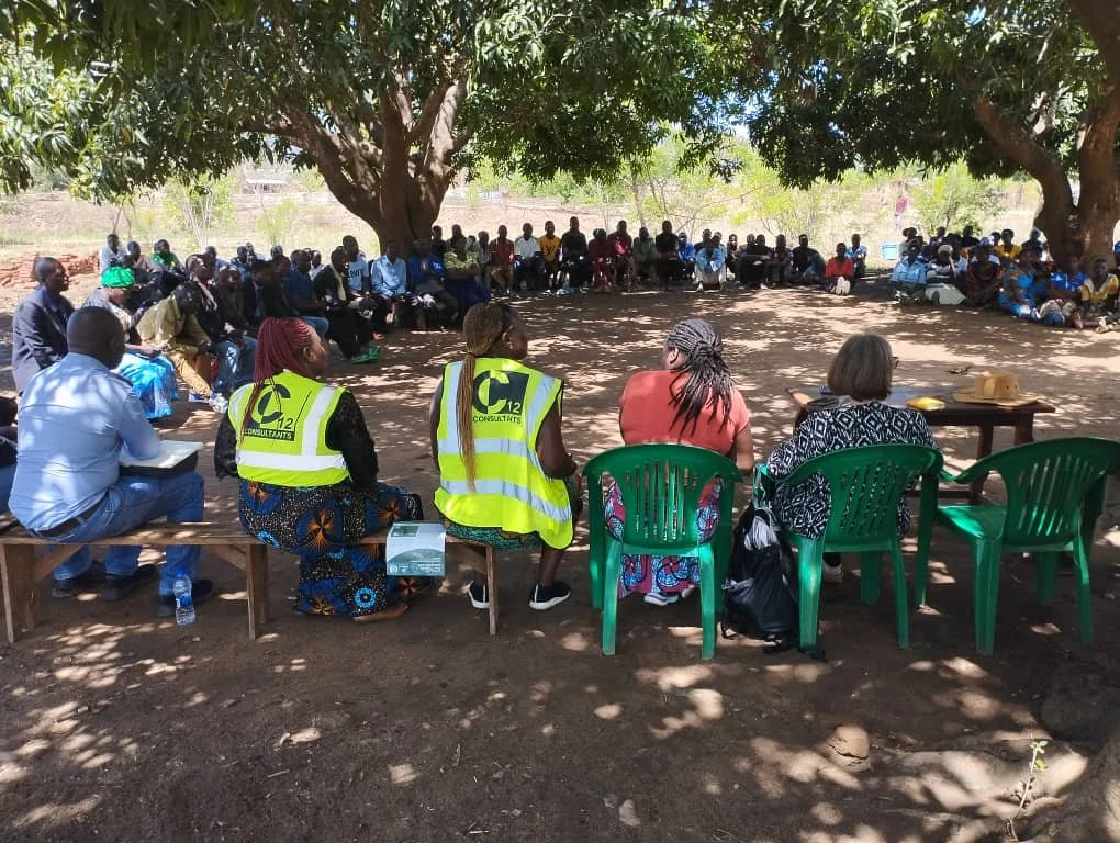 A large group of people gathered in a wide circle for a community meeting under the shade of large trees. Several facilitators in the foreground, two wearing bright yellow safety vests, sit on a wooden bench and green plastic chairs facing the crowd.