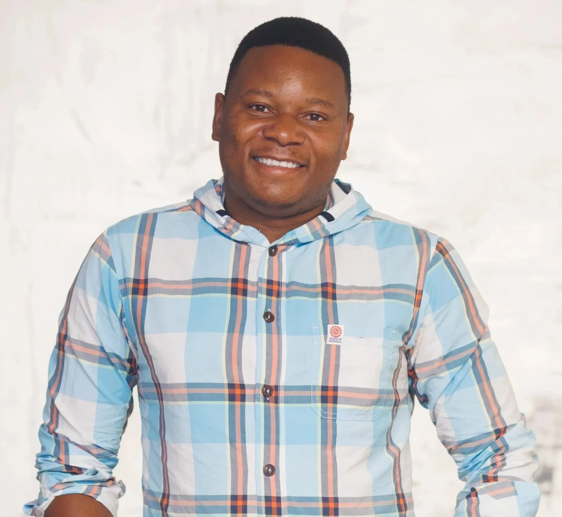 A head-and-shoulders portrait of William Rozario, smiling with a short haircut. He is wearing a light blue, white, and orange plaid button-down shirt with a hood. He stands against a neutral, slightly textured off-white background.