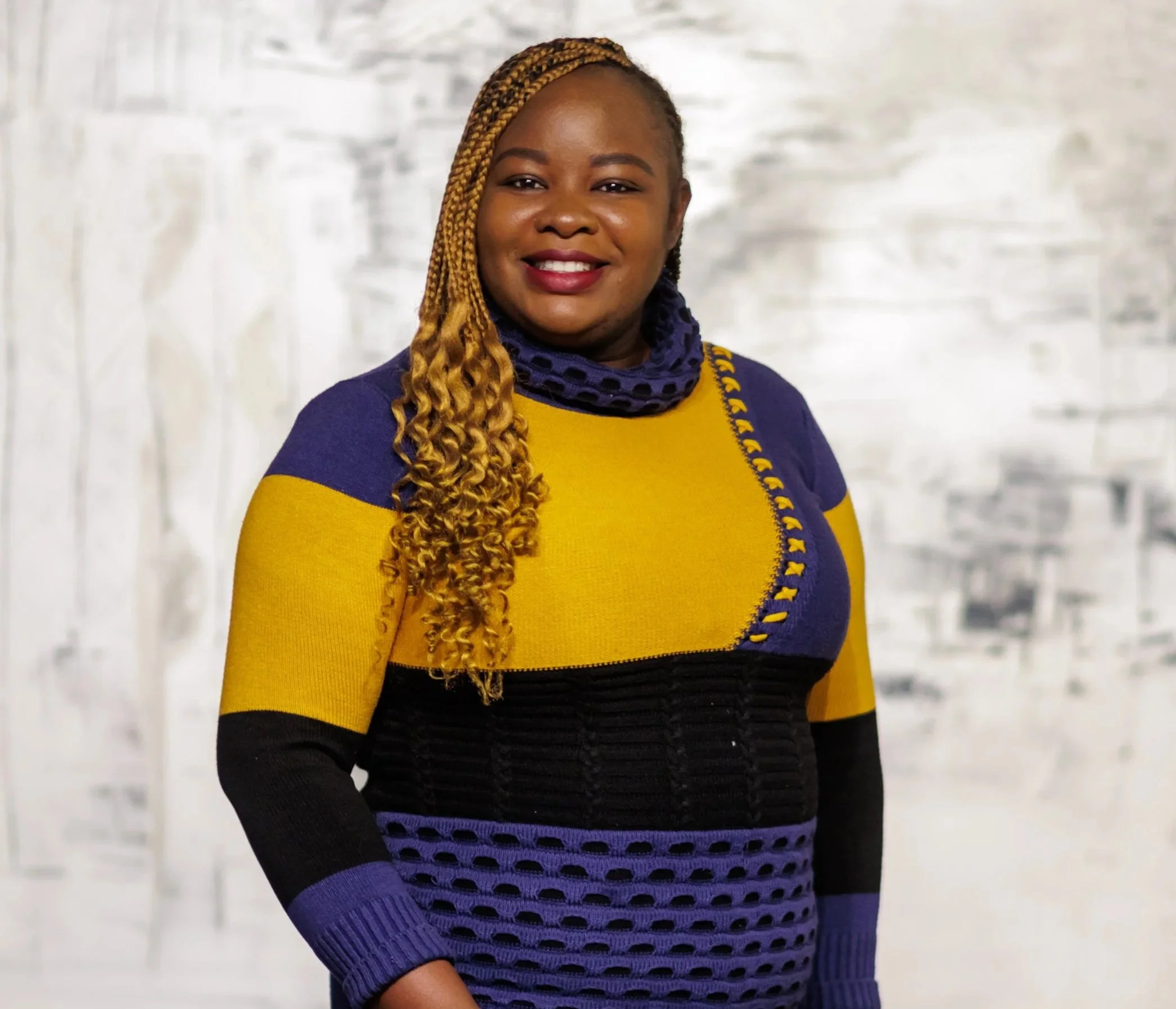 Gemini said A smiling woman with blonde braided hair wearing a vibrant, color-blocked sweater in yellow, blue, and black.