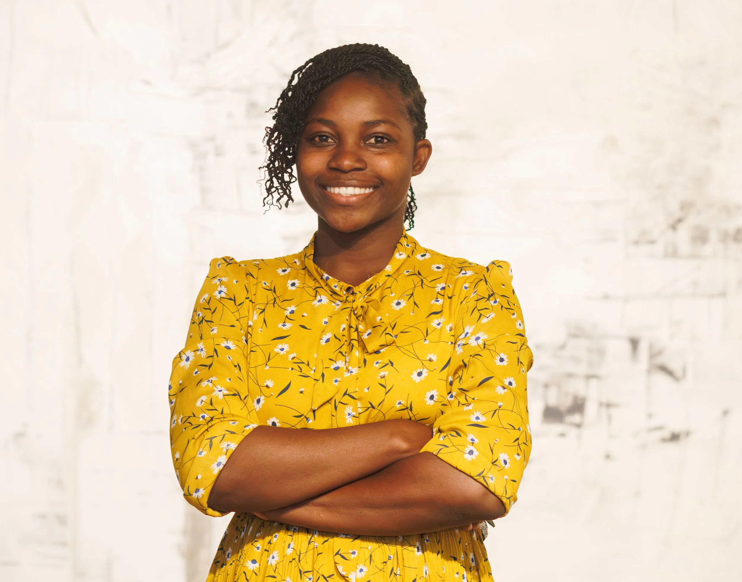 A smiling woman with her arms crossed, wearing a yellow floral-patterned dress and her hair styled in side-swept twists.