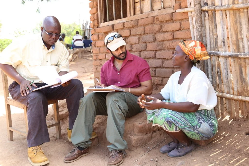 Two men conducting an interview with a woman outside a brick building. One man sits on a wooden chair reviewing documents, the other sits on a low brick ledge taking notes, and the woman is seated on the ground speaking to them.
