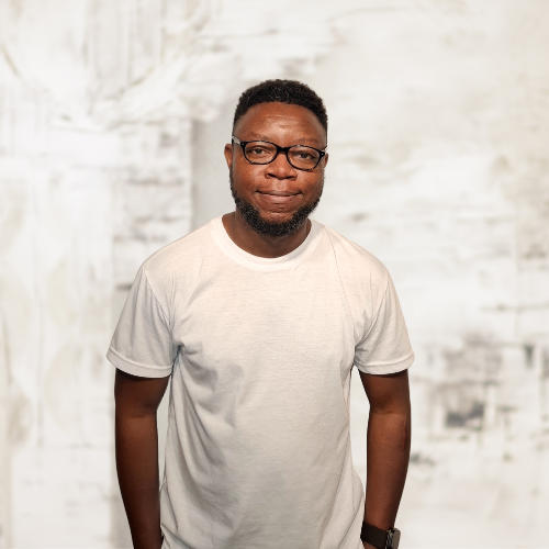A medium-close up photograph of a smiling, dark-skinned man with short hair, black-rimmed glasses, and a short beard, wearing a white t-shirt, standing with hands in pockets against a blurred light-grey background.