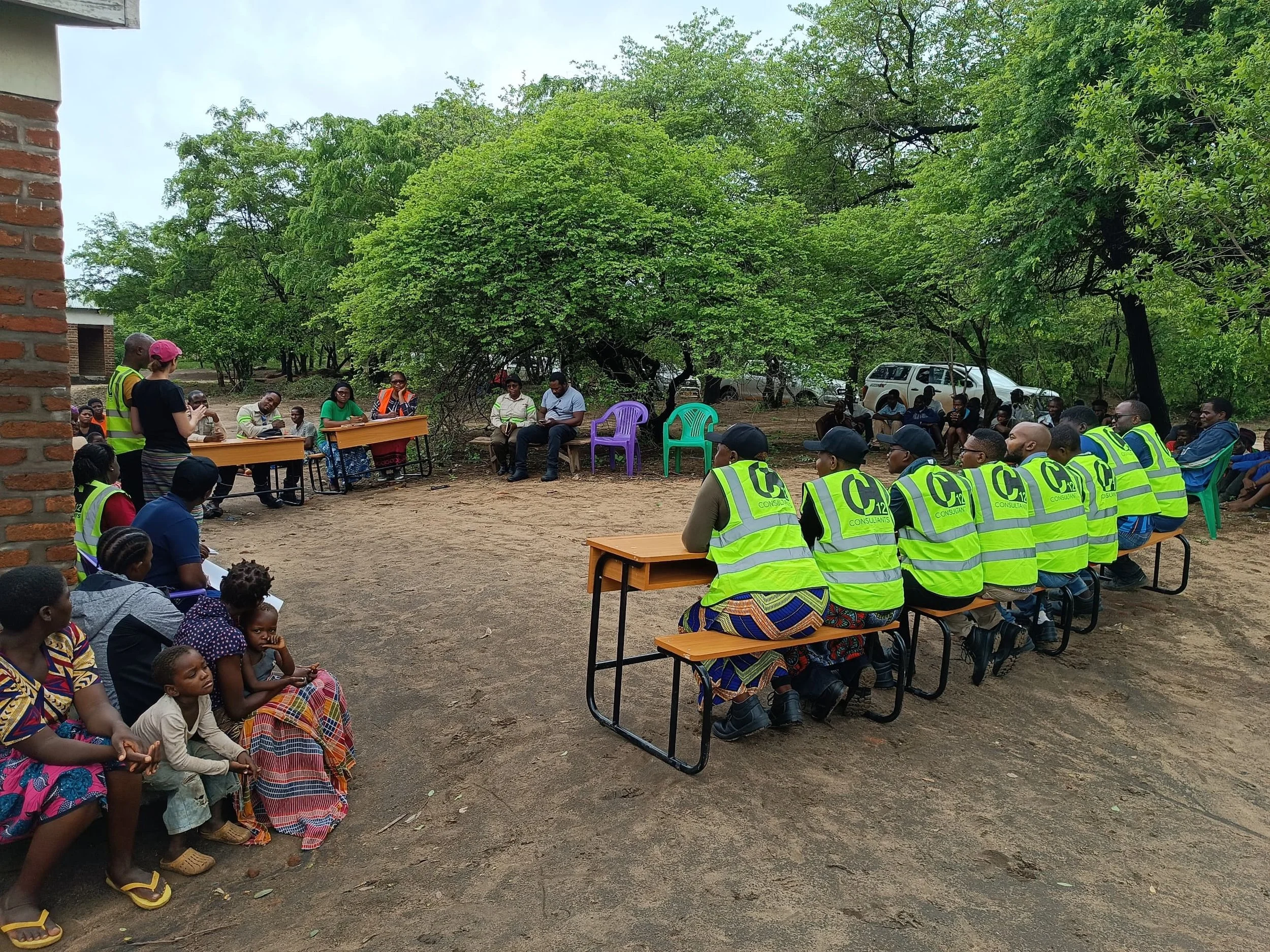 An outdoor community meeting in a wooded clearing. Facilitators and field workers in neon safety vests sit at a desk facing a large group of seated community members.