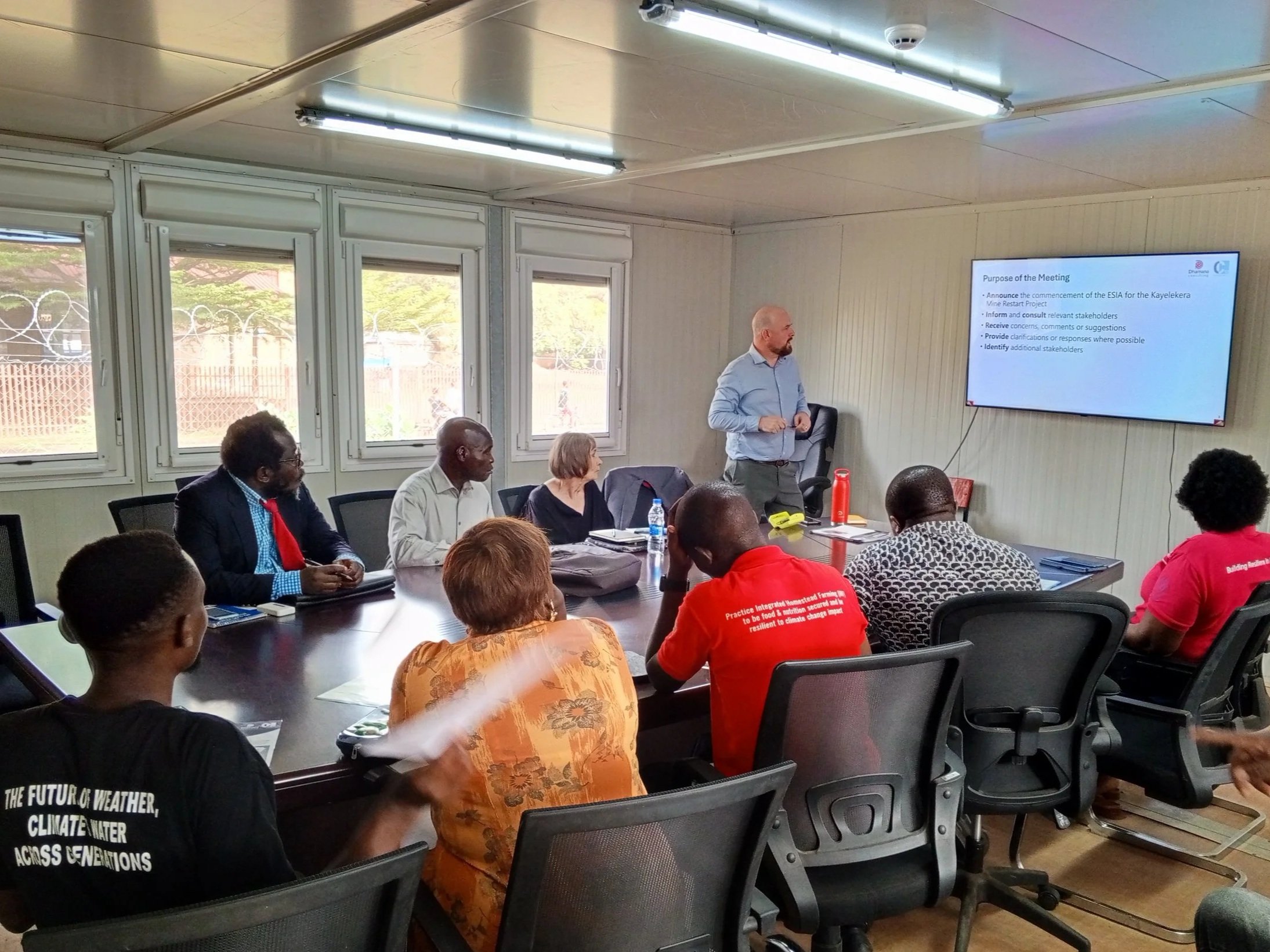An indoor office setting where a professional meeting is taking place. A man in a patterned shirt sits at a large desk, gesturing as he speaks to a group of seven colleagues seated in chairs along the wall.