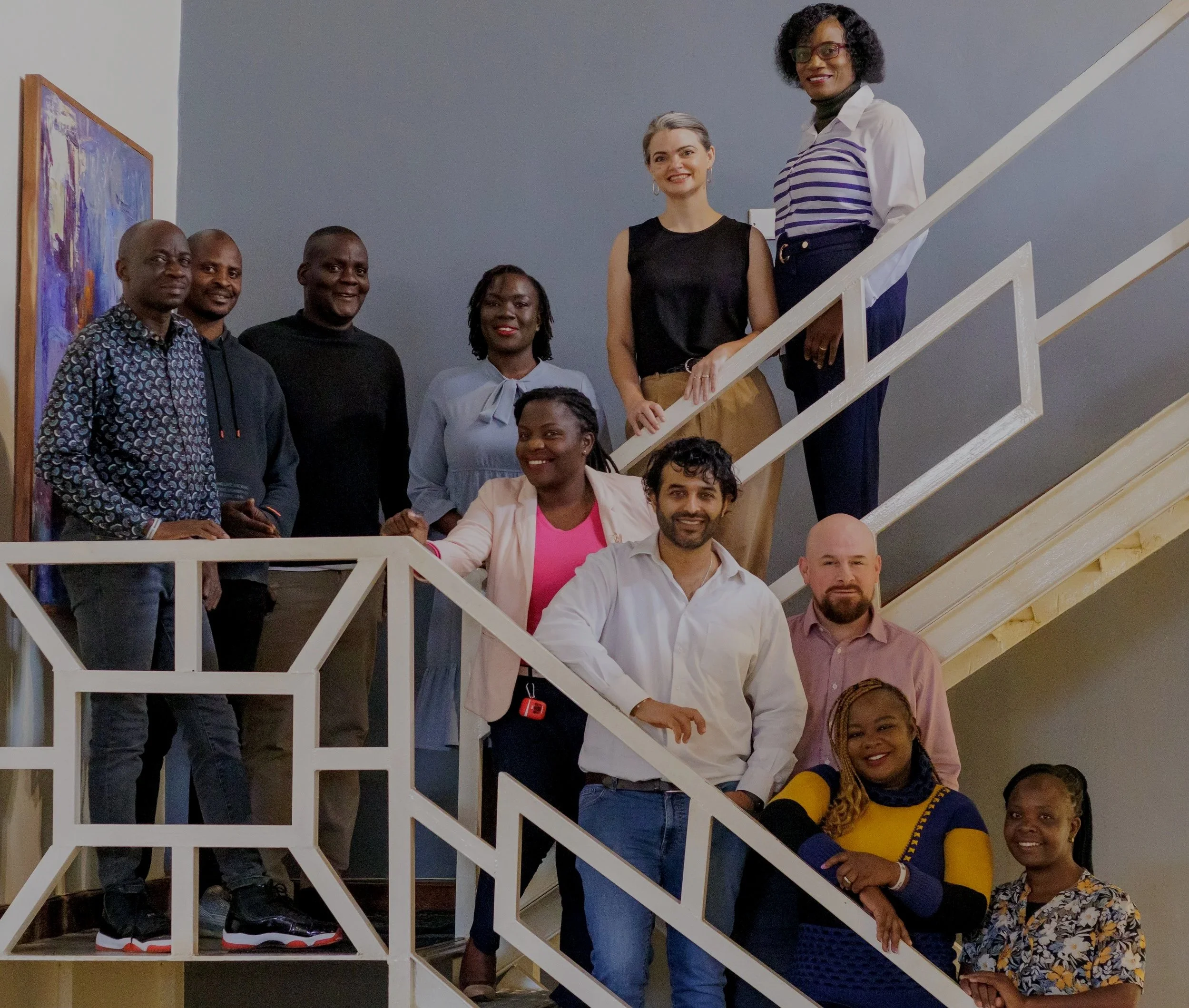 A diverse group of eleven professionals posing for a team photo on a modern white staircase. The team members are smiling and arranged at different levels of the stairs against a neutral blue and white wall.
