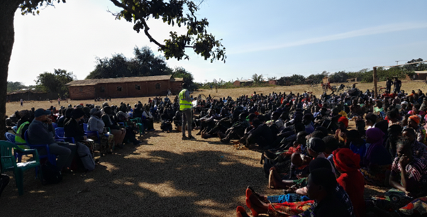 A wide-angle shot of a large community gathering outdoors. A facilitator wearing a bright neon-yellow safety vest stands in the center, addressing a large crowd of people seated on the ground in a sunlit clearing.