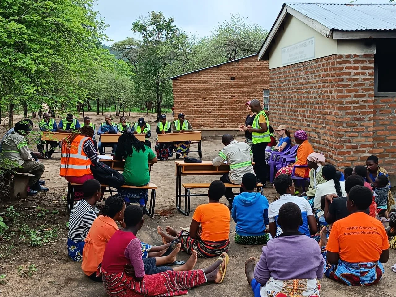 An outdoor meeting where a man in a neon safety vest stands and addresses a large group of community members seated on the ground. A row of field workers, also in safety vests, sit at wooden desks behind him